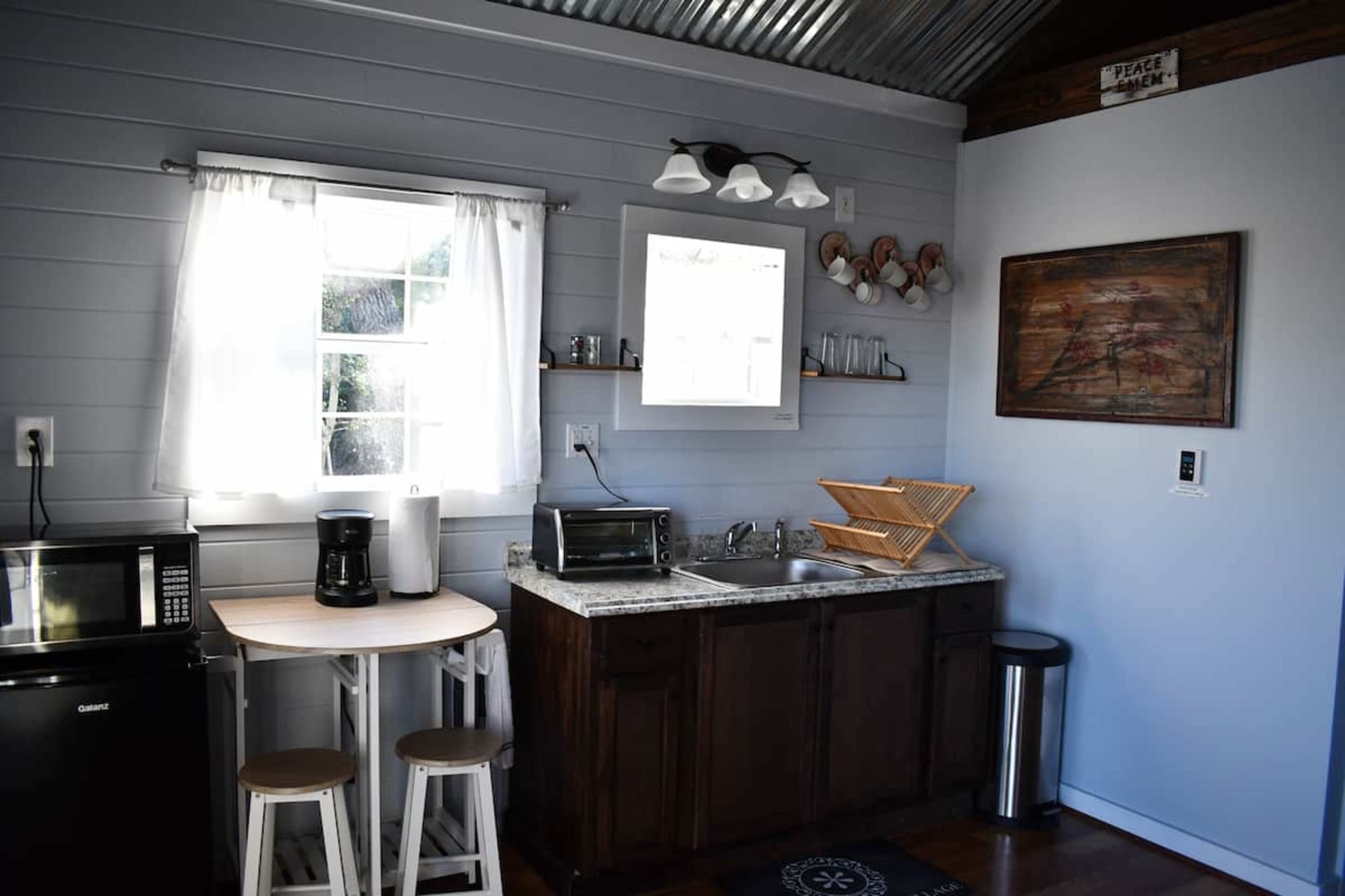 A compact kitchen featuring a countertop with a sink, a microwave, a coffee maker, and a small table with two stools, all against a backdrop of light blue walls and metal accents.