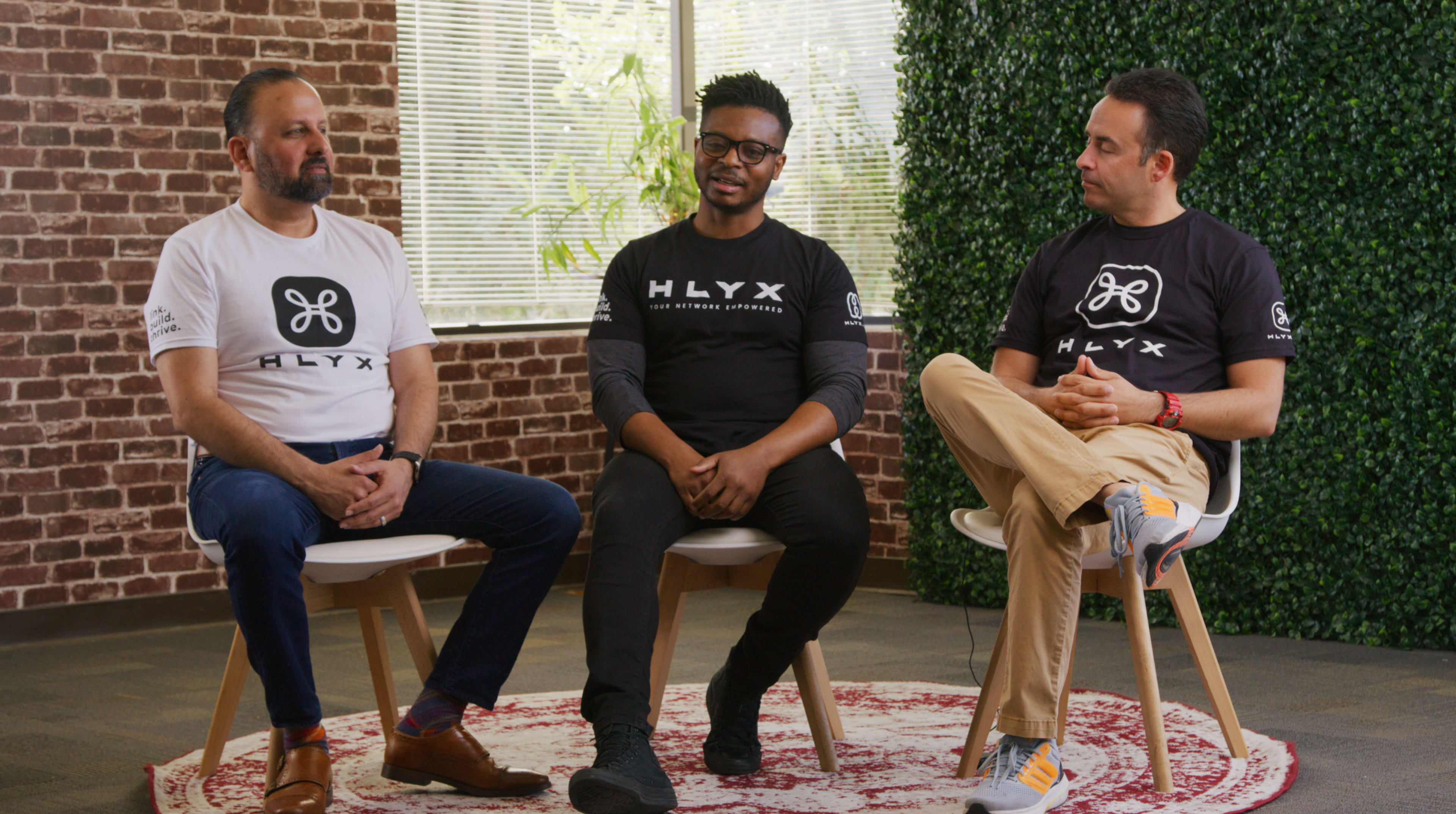 Three men sit on chairs in a well-lit room, wearing matching branded T-shirts, engaged in conversation.
