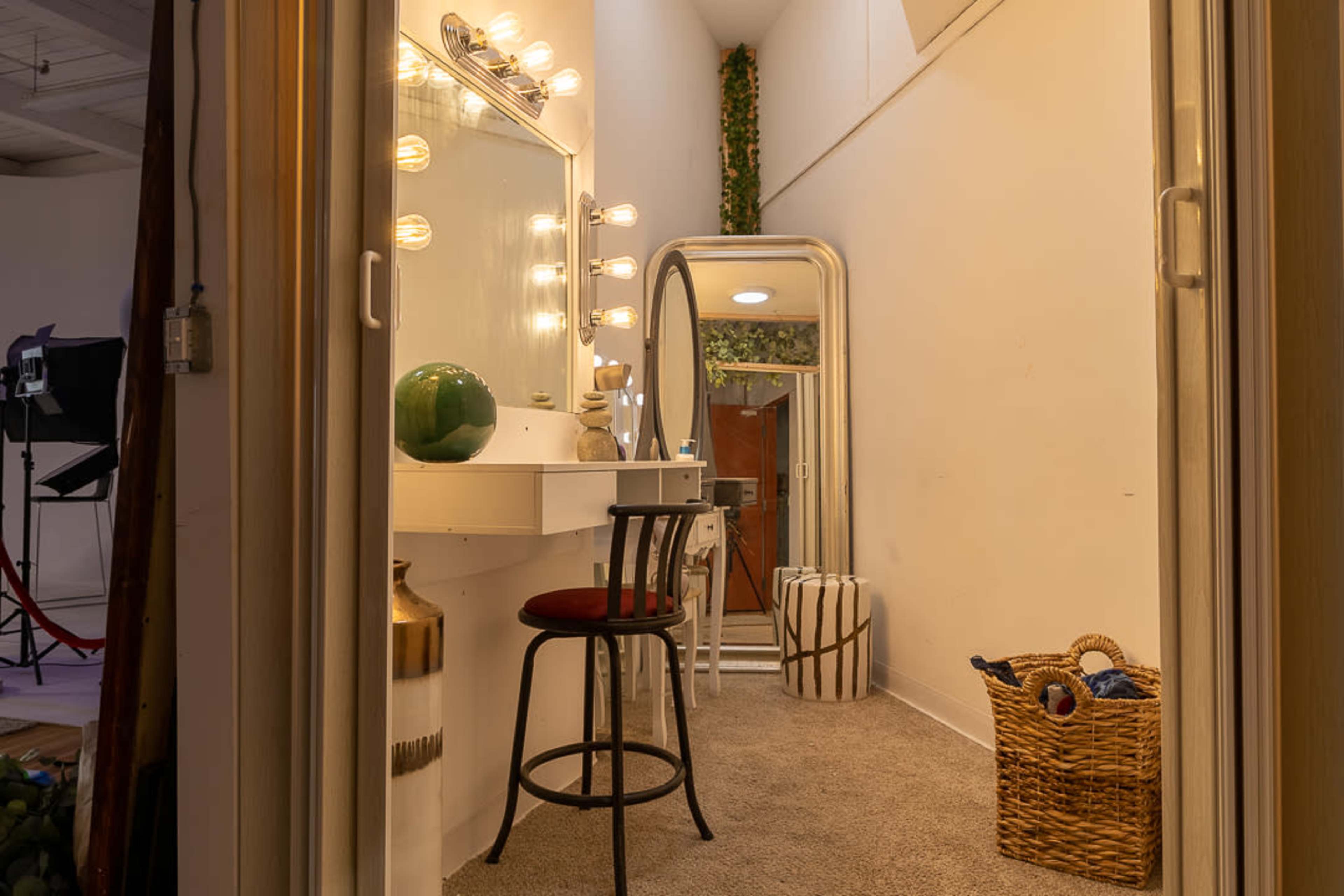 The image shows a well-lit dressing area featuring a vanity with lights, a mirror, a chair, and decorative items in a cozy hallway.