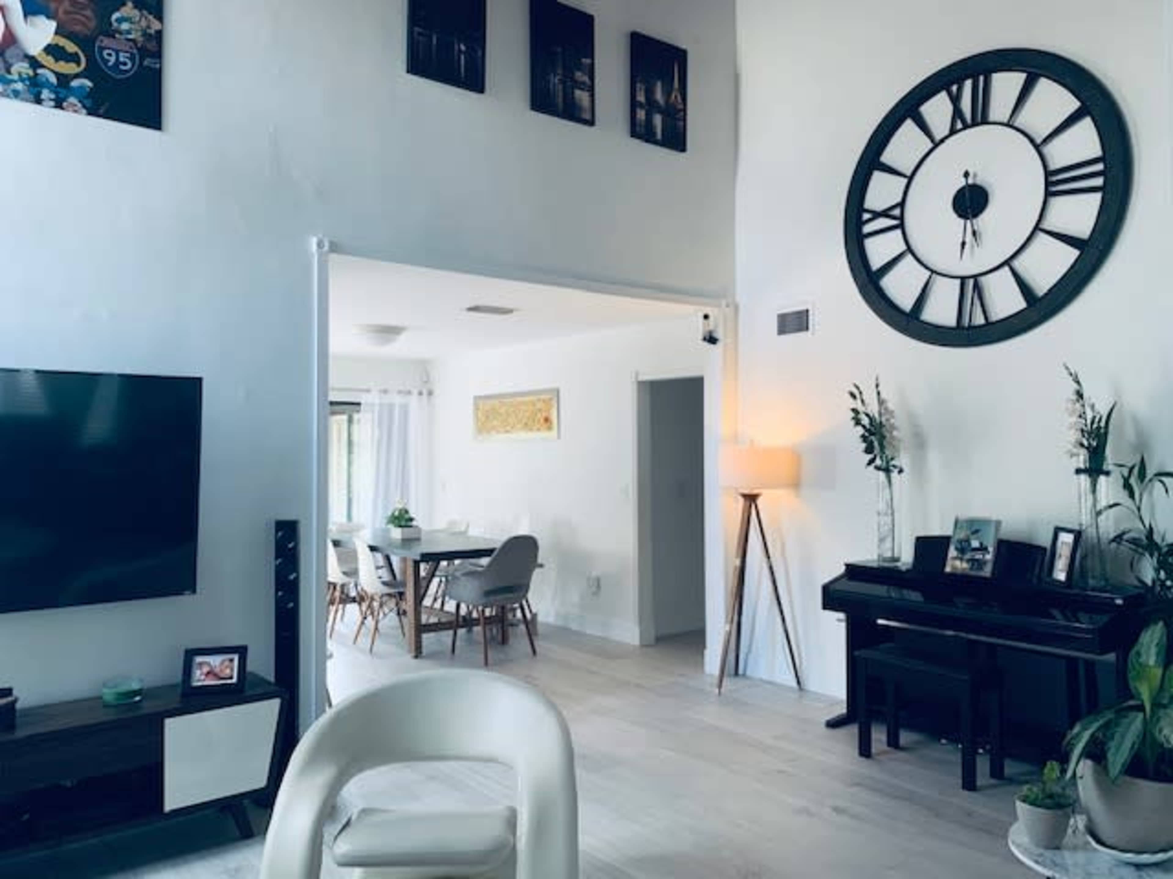 The image shows a modern living room featuring a large clock on the wall, a piano, a television, and a dining area visible through an adjoining space.
