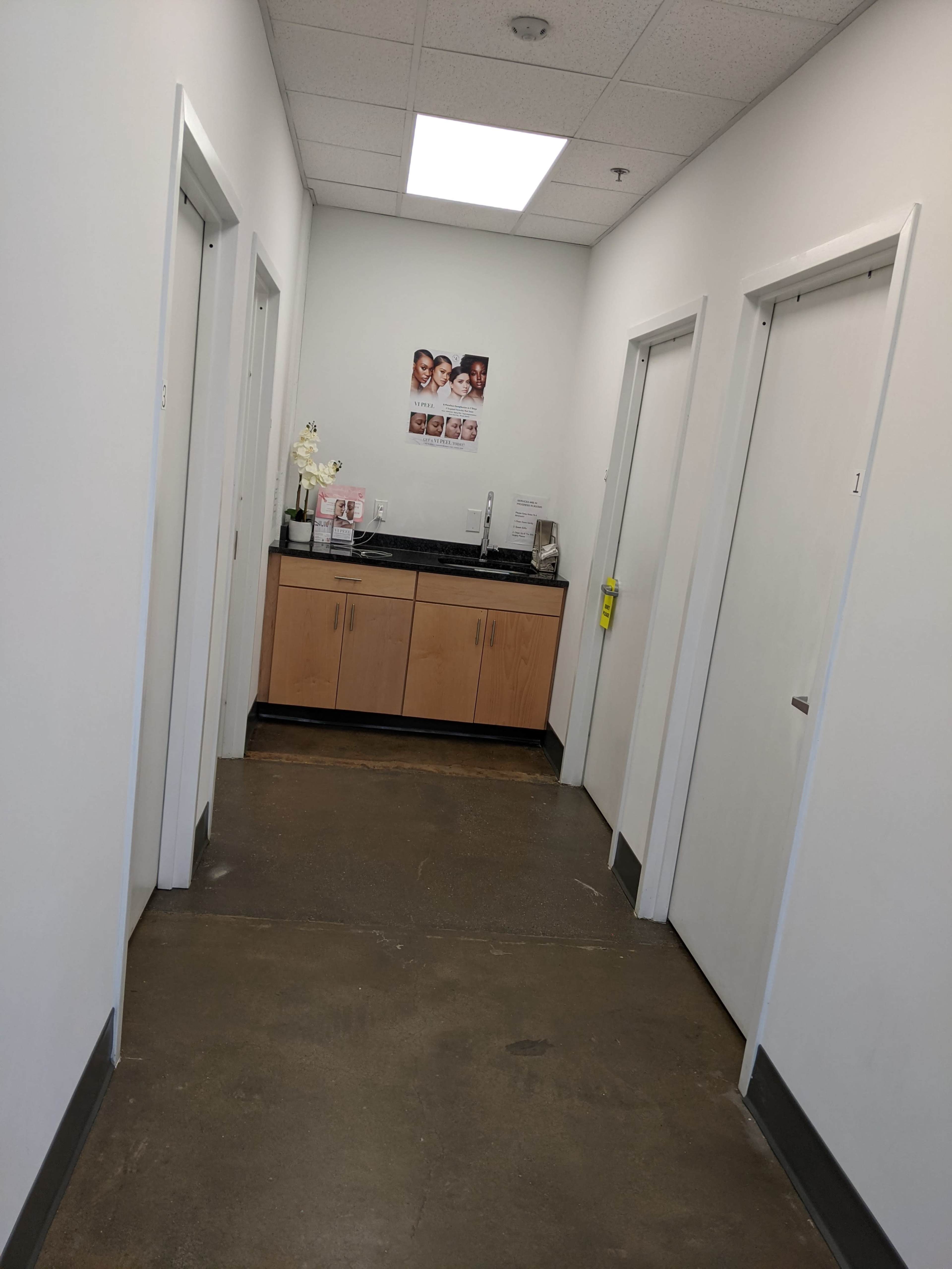 A narrow hallway features three doors on each side, a countertop with a dispenser, and a poster on the wall.