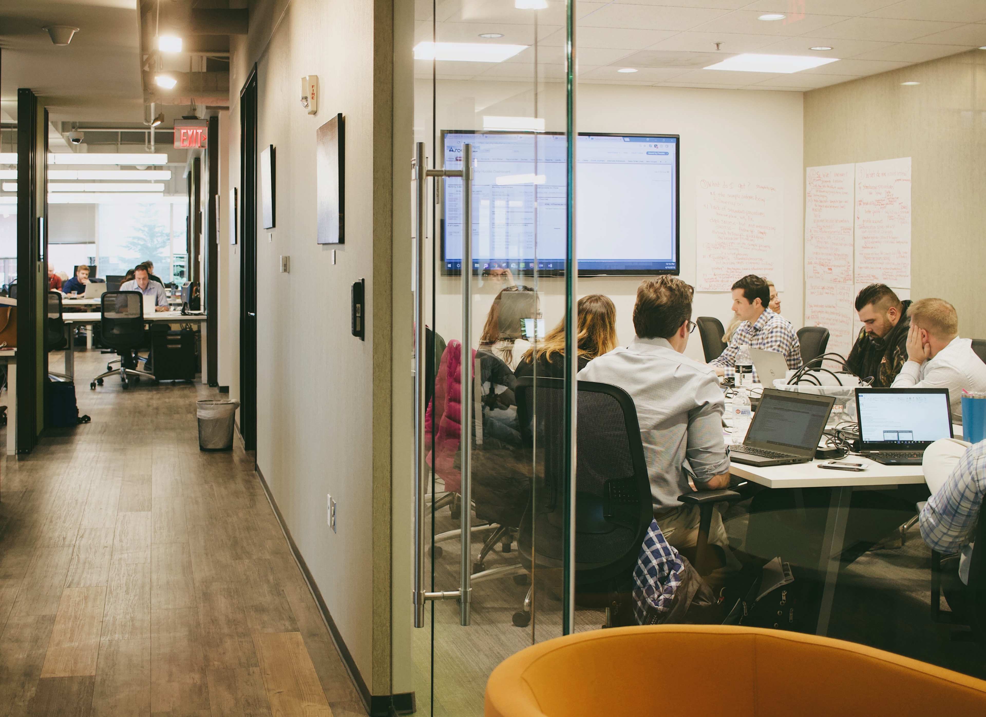 A group of people is seated around a conference table in a modern office, with a large screen displaying information in the background.