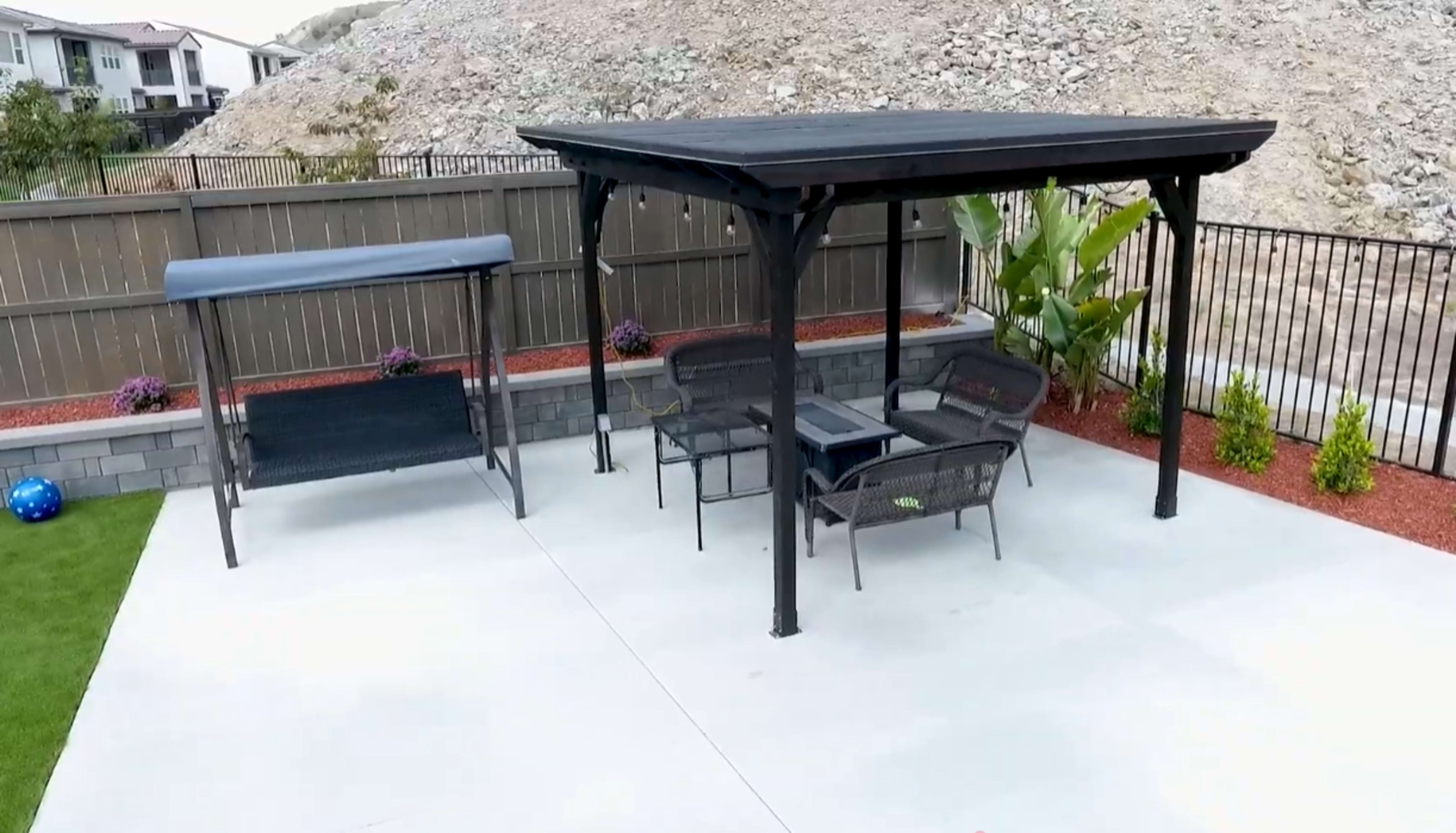 The image shows a patio area featuring a black gazebo, a seating arrangement with chairs and a table, and a swing set in a landscaped backyard.