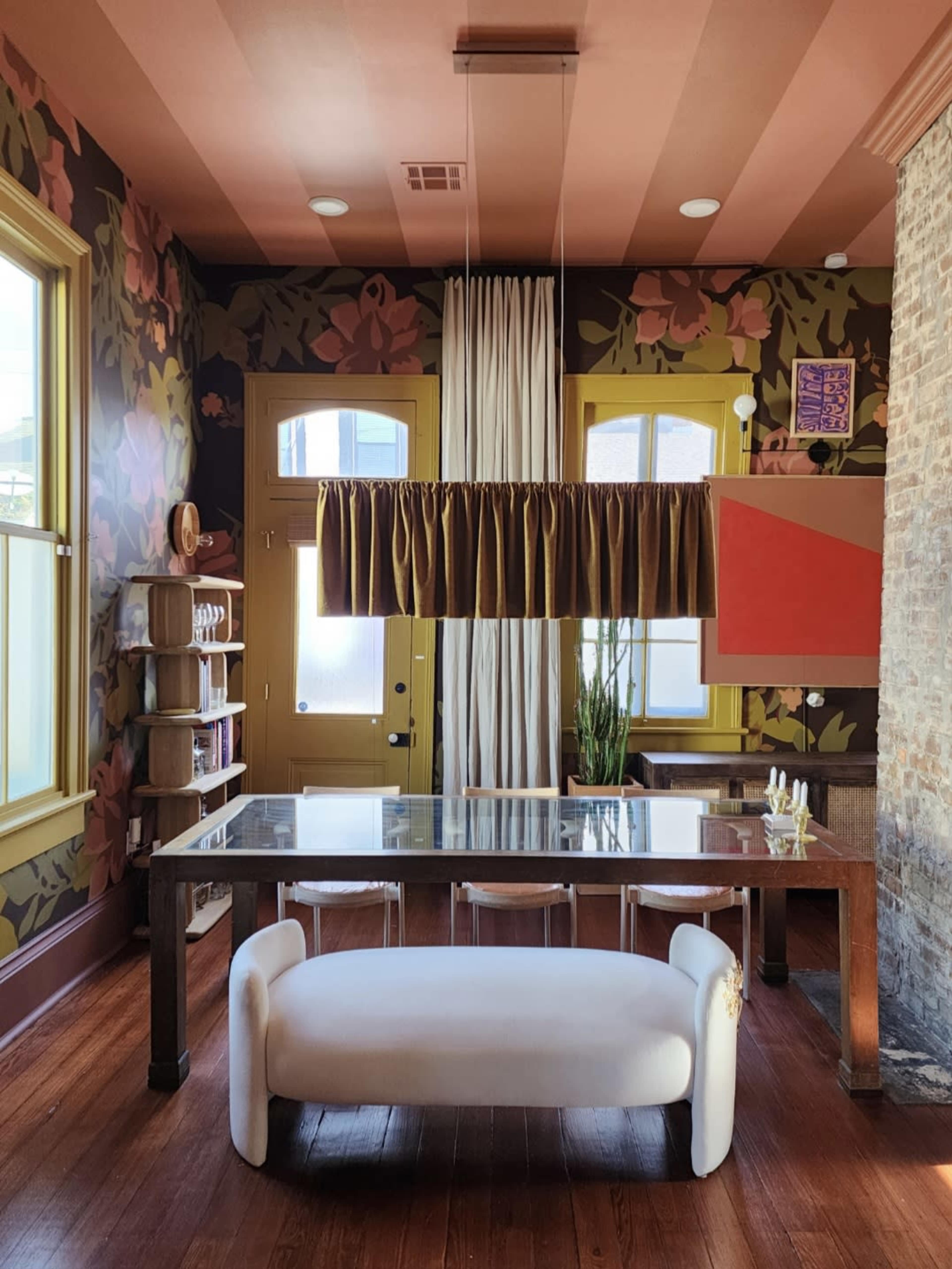 The image shows a dining area with a long glass table surrounded by clear chairs, alongside floral wallpaper, a large pendant light, and a stone wall.