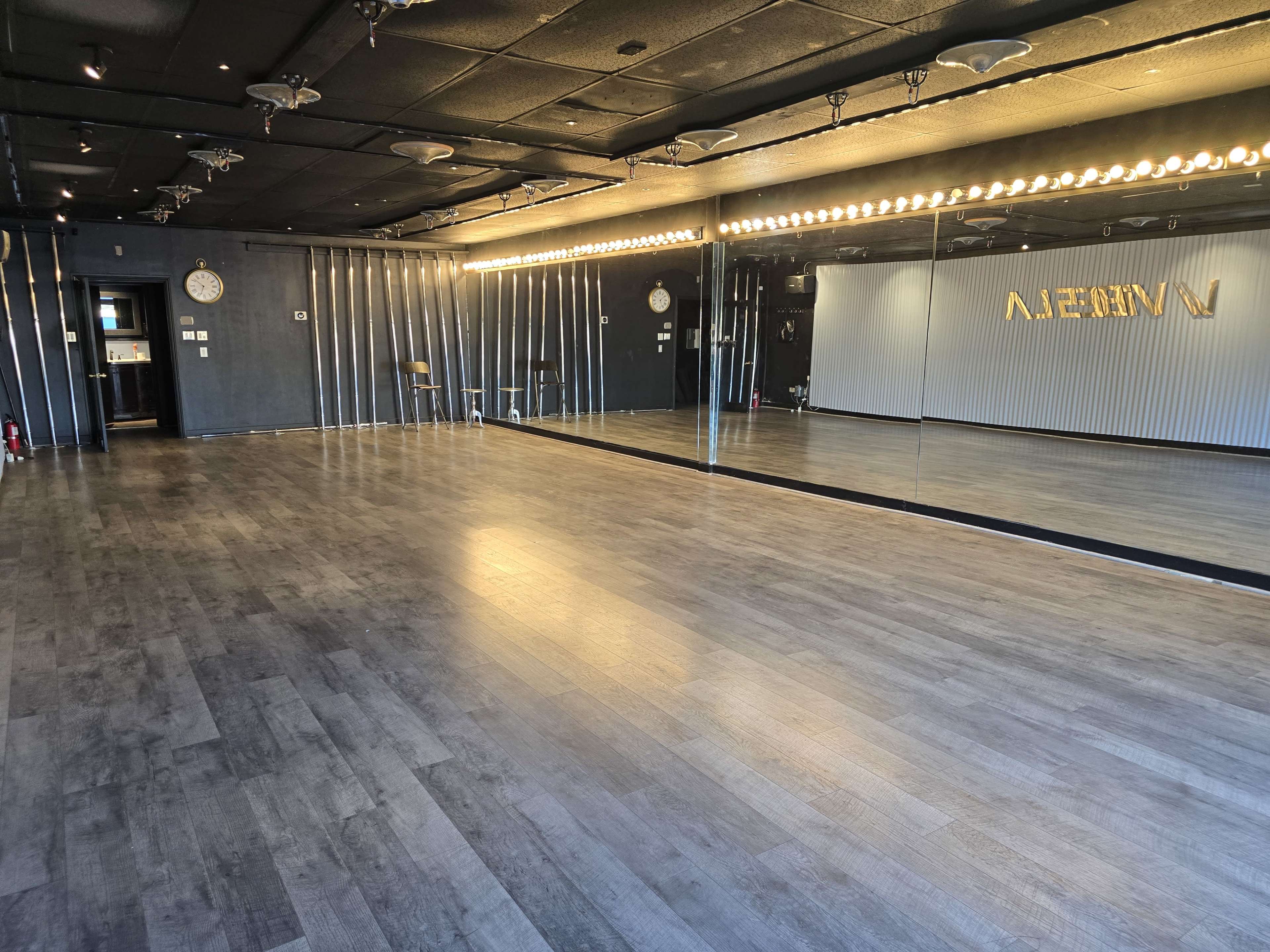 The image shows an empty dance studio with wooden flooring, large mirrors on one wall, and dim lighting fixtures along the ceiling.