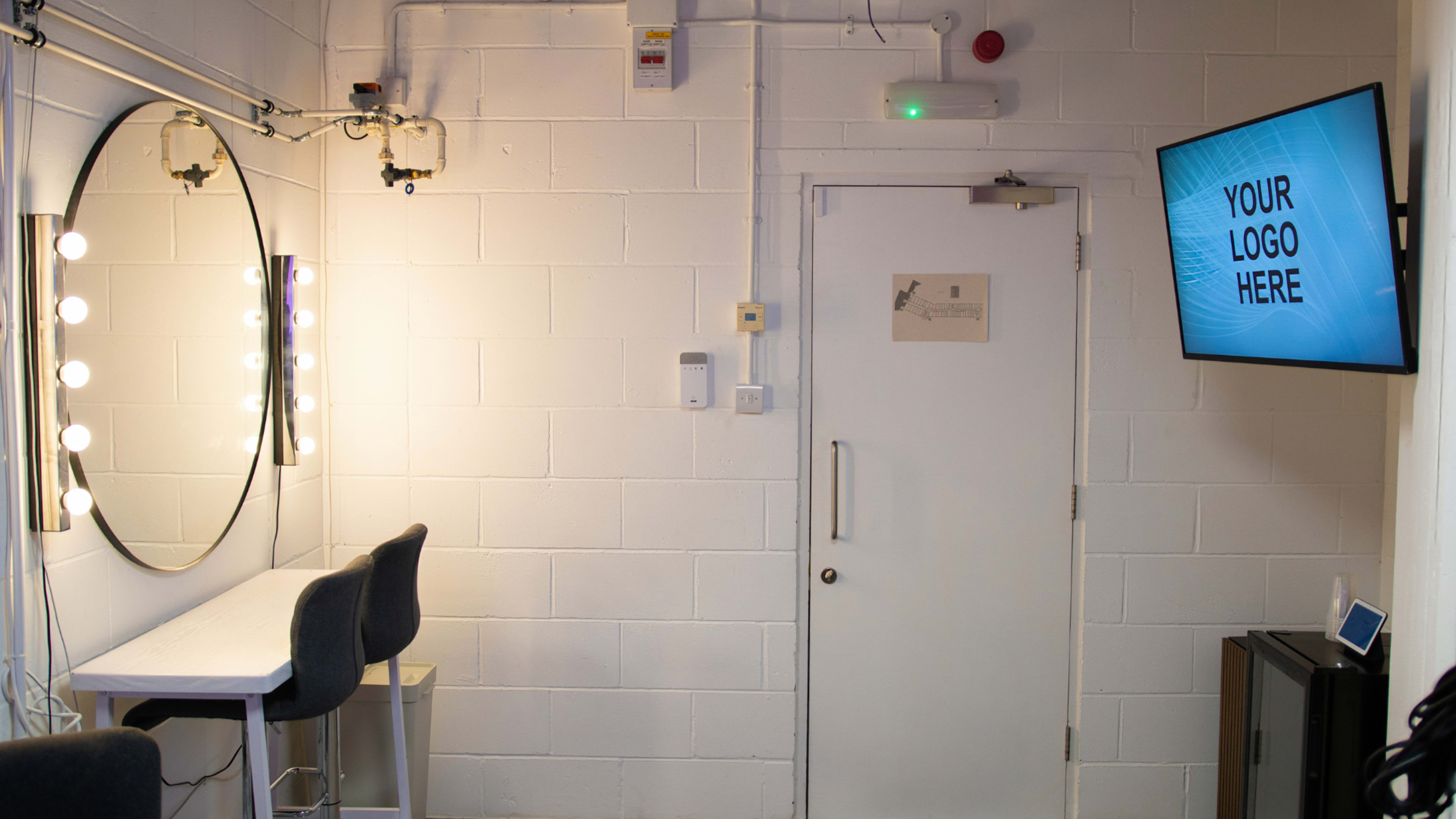 The image shows a well-lit dressing room with a circular mirror and lights, a small table with two stools, and a door leading to another area, along with a television displaying a placeholder for a logo.