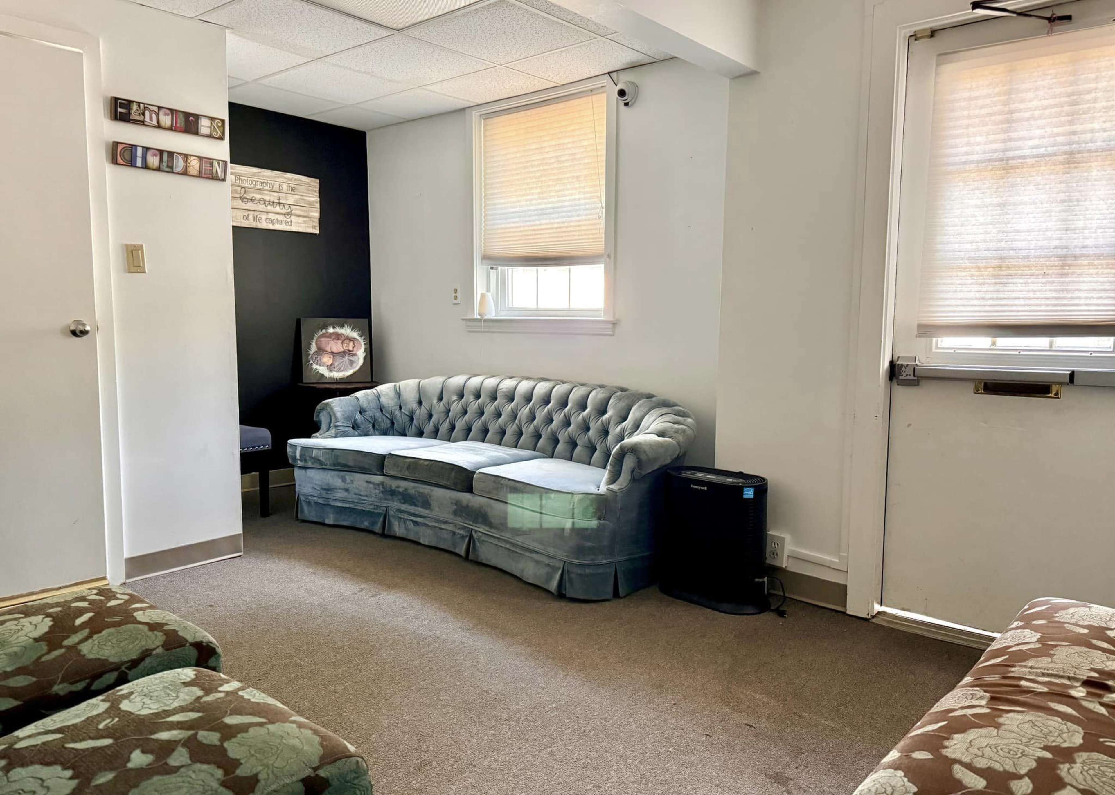 The image shows a room with a tufted blue couch, a small table on the left wall, and a door leading outside.