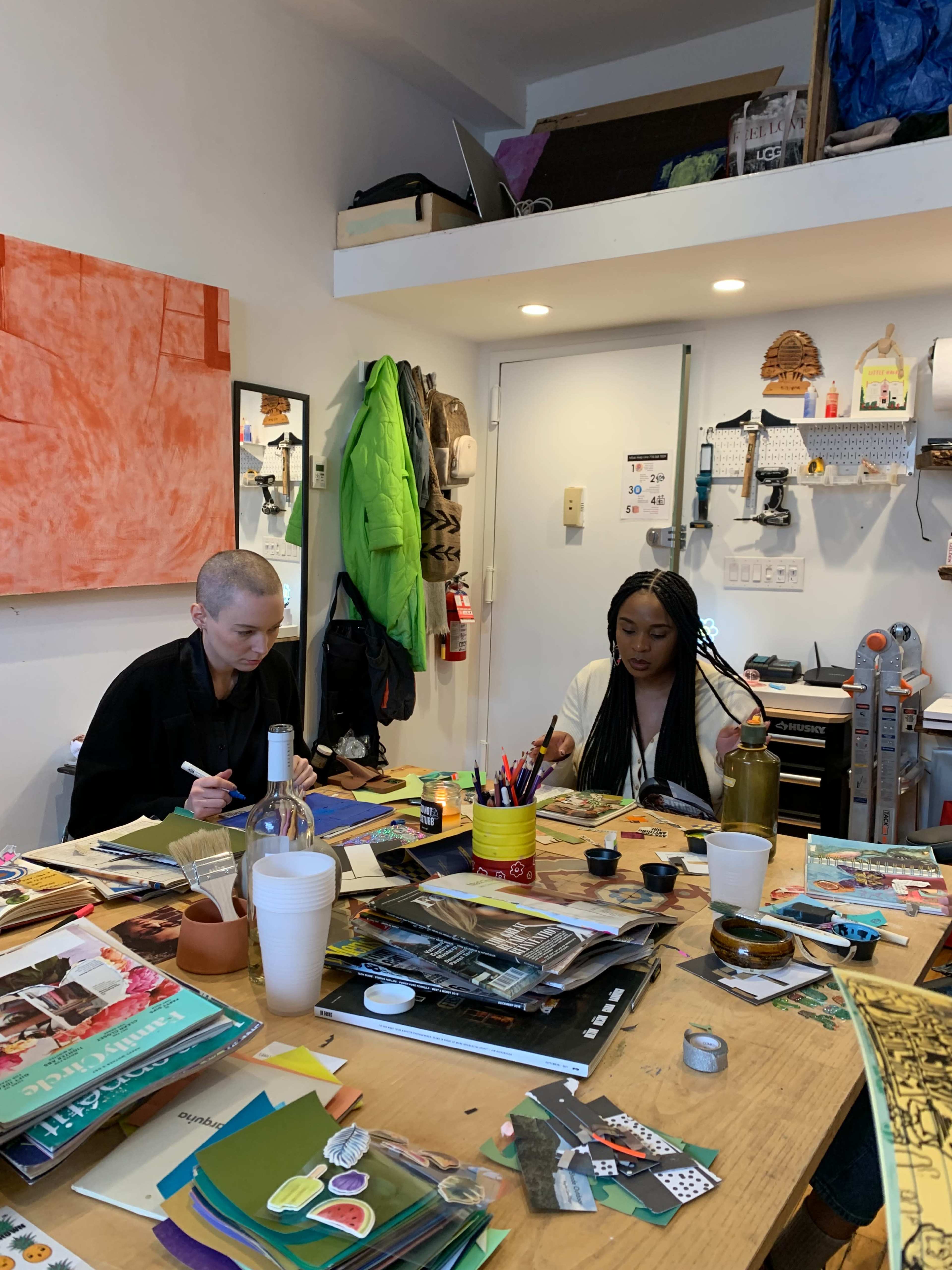 Two people are sitting at a table covered with art supplies and materials in a studio environment.