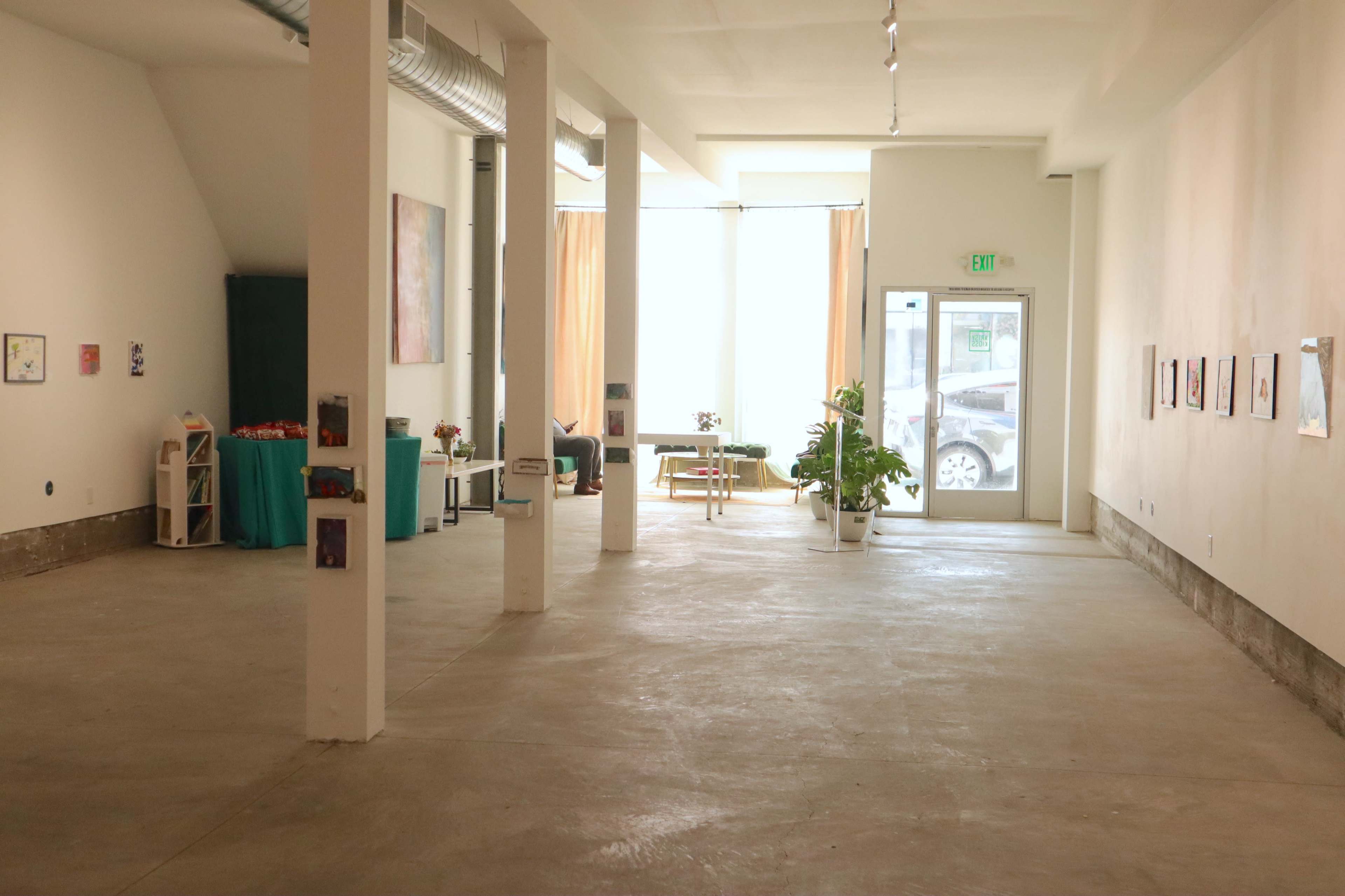 The image shows an empty art gallery with large windows allowing natural light, featuring minimal furniture and framed artworks on the walls.