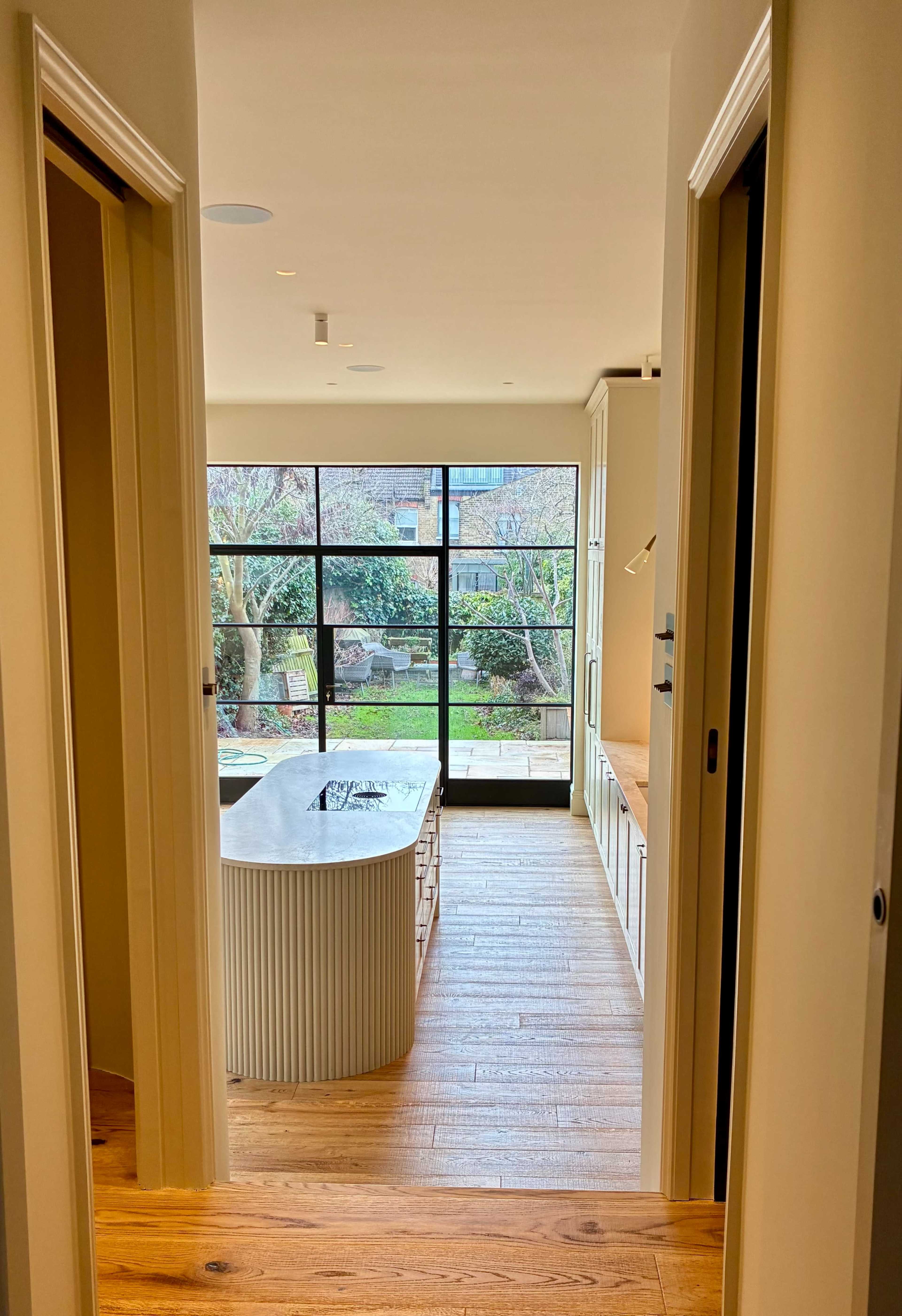 The image shows a view of a modern kitchen with a large island, framed by two doorways and overlooking a garden through large glass windows.