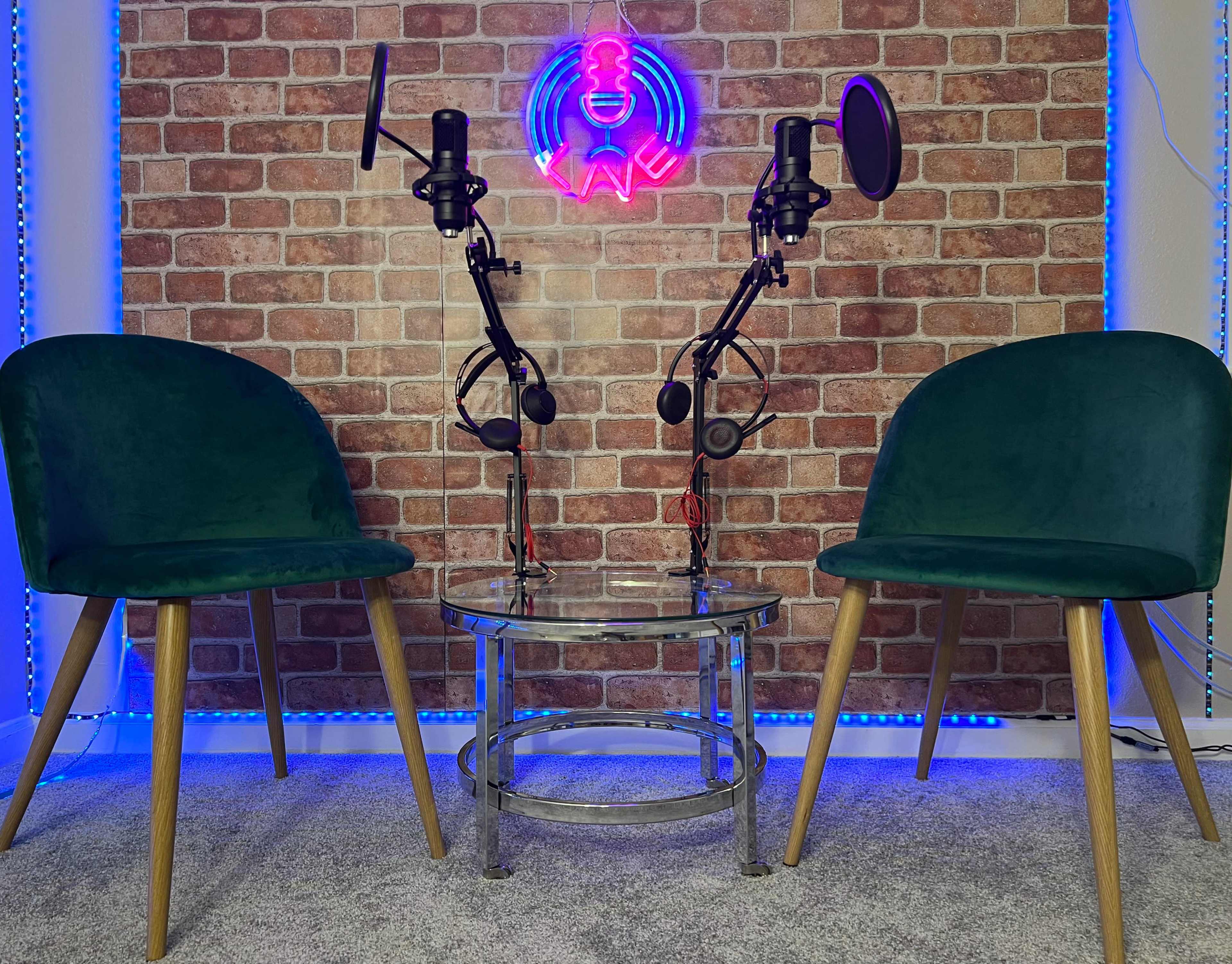 The image shows two green chairs on either side of a glass table, with two microphone stands positioned above the table against a brick-patterned wall featuring a neon sign.