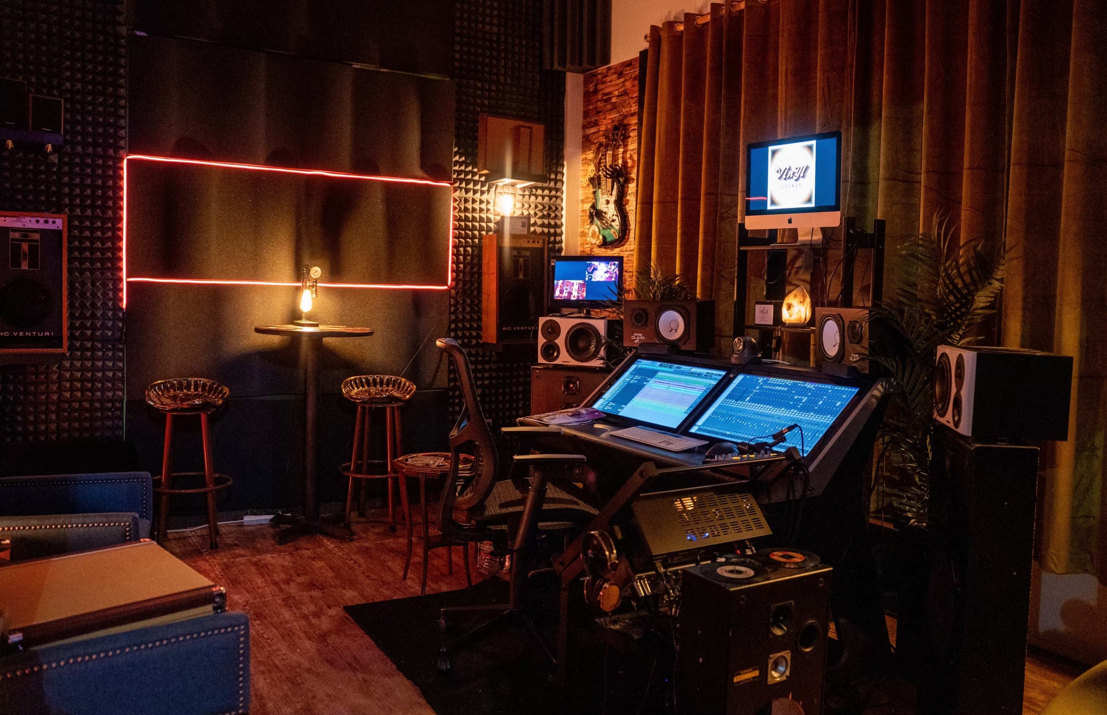 A music studio featuring a large mixing console, speakers, and a cozy seating area with bar stools against a backdrop of acoustic panels.