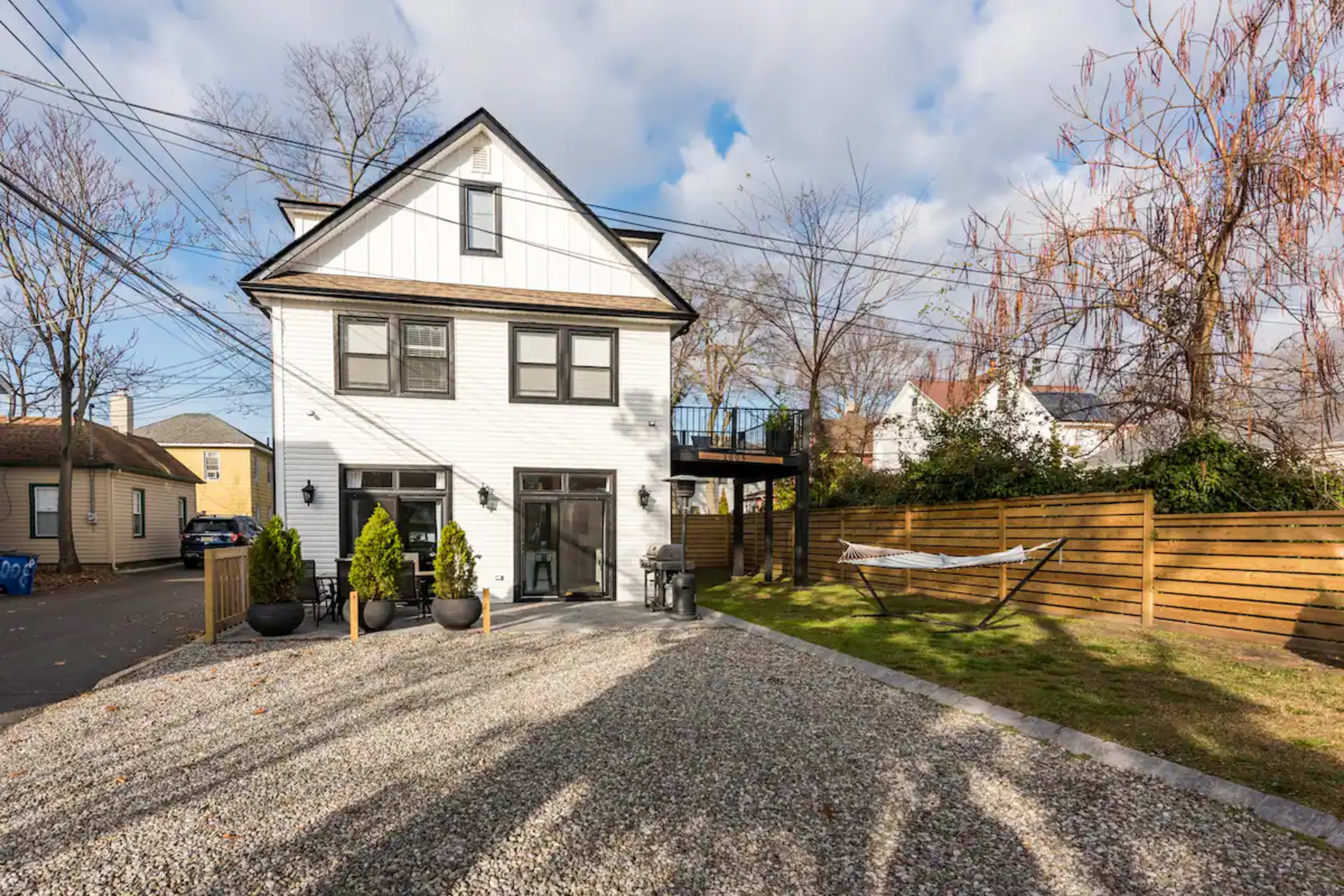 A three-story white house with large windows is situated on a gravel driveway surrounded by a wooden fence and a hammock in the yard.