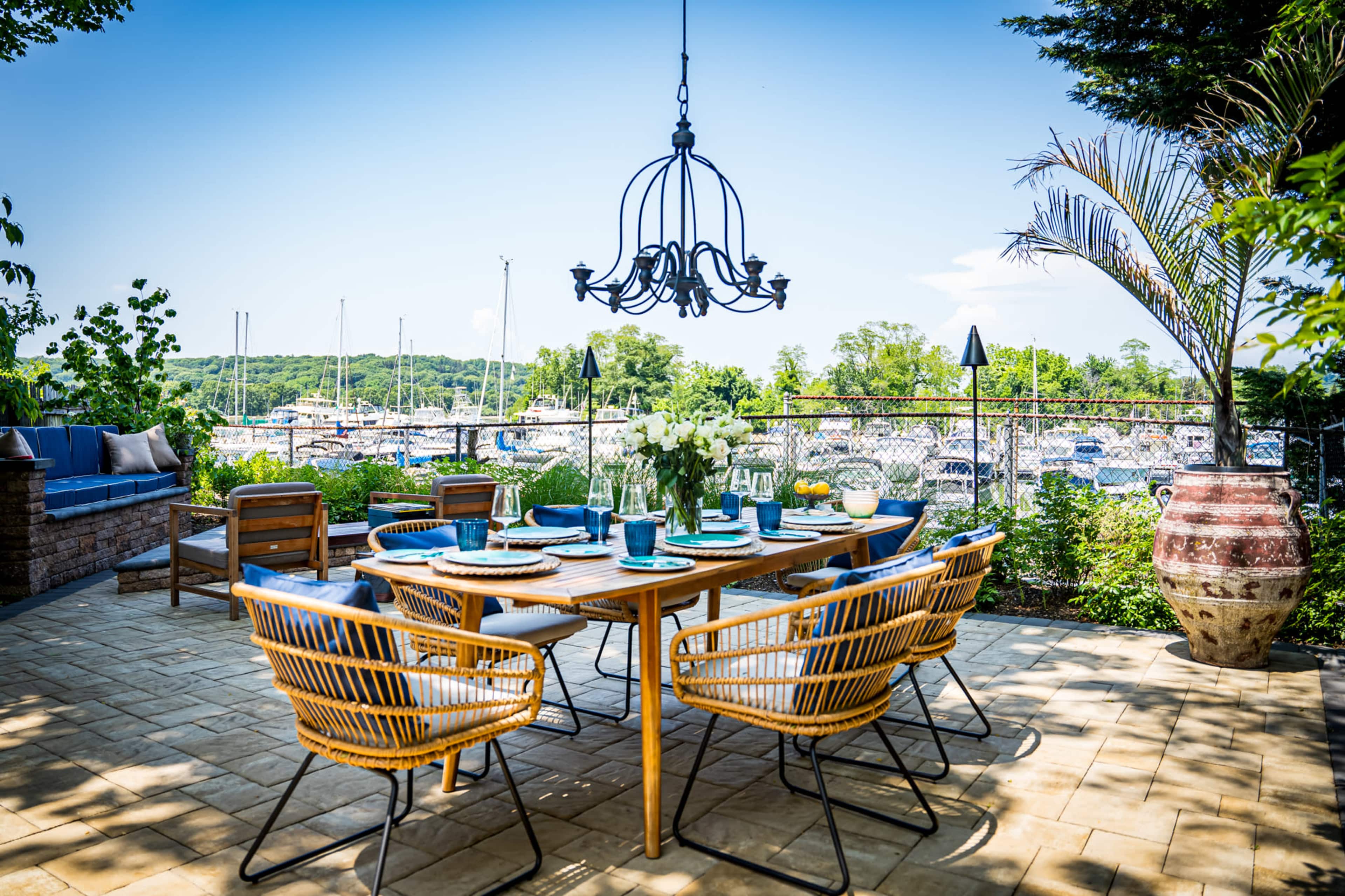 A dining area with a large table set for a meal overlooks a marina with boats and lush greenery in the background.