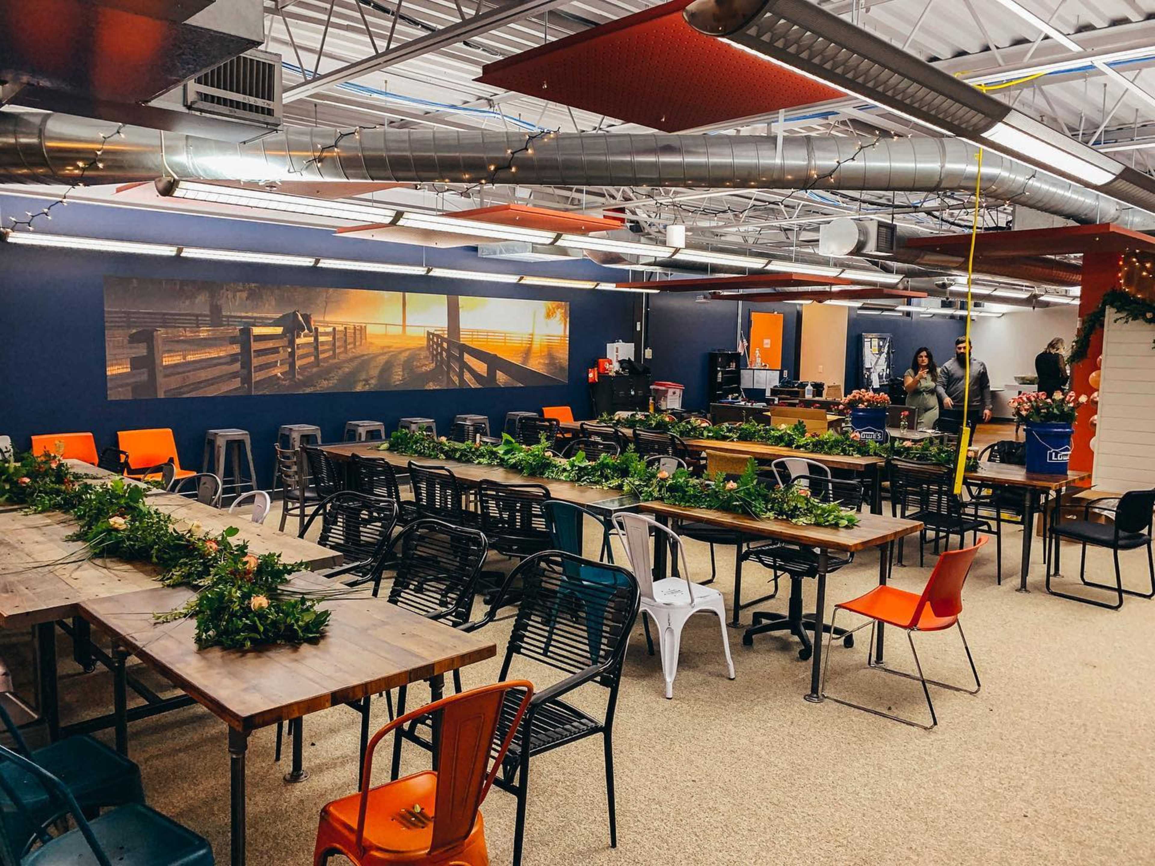 A large, open room is filled with long communal tables and chairs, featuring greenery decor on the tables and a wall mural of a scenic pathway.