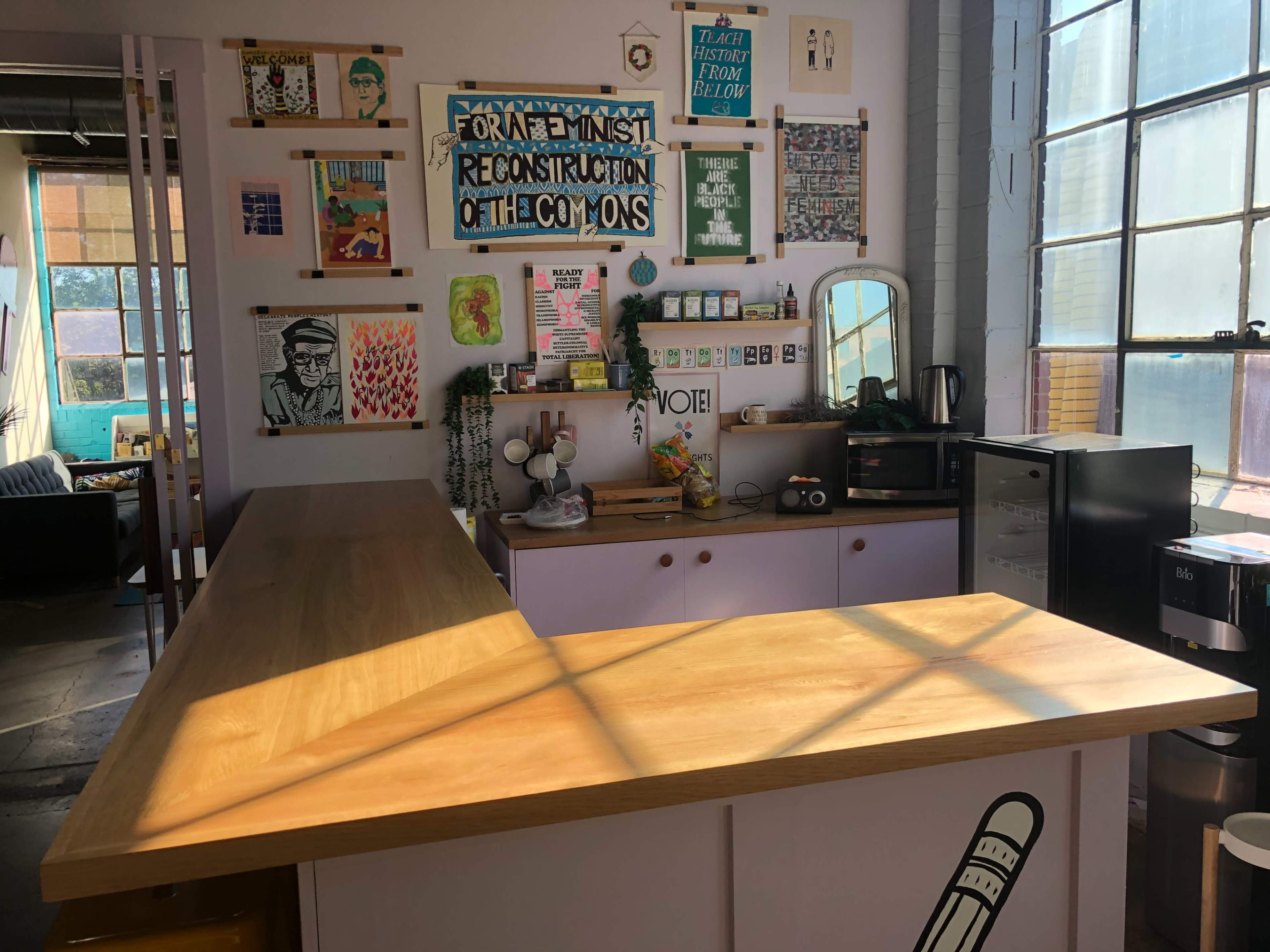 A brightly lit coffee shop interior with a wooden counter, colorful artwork on the walls, and a small refrigerator.