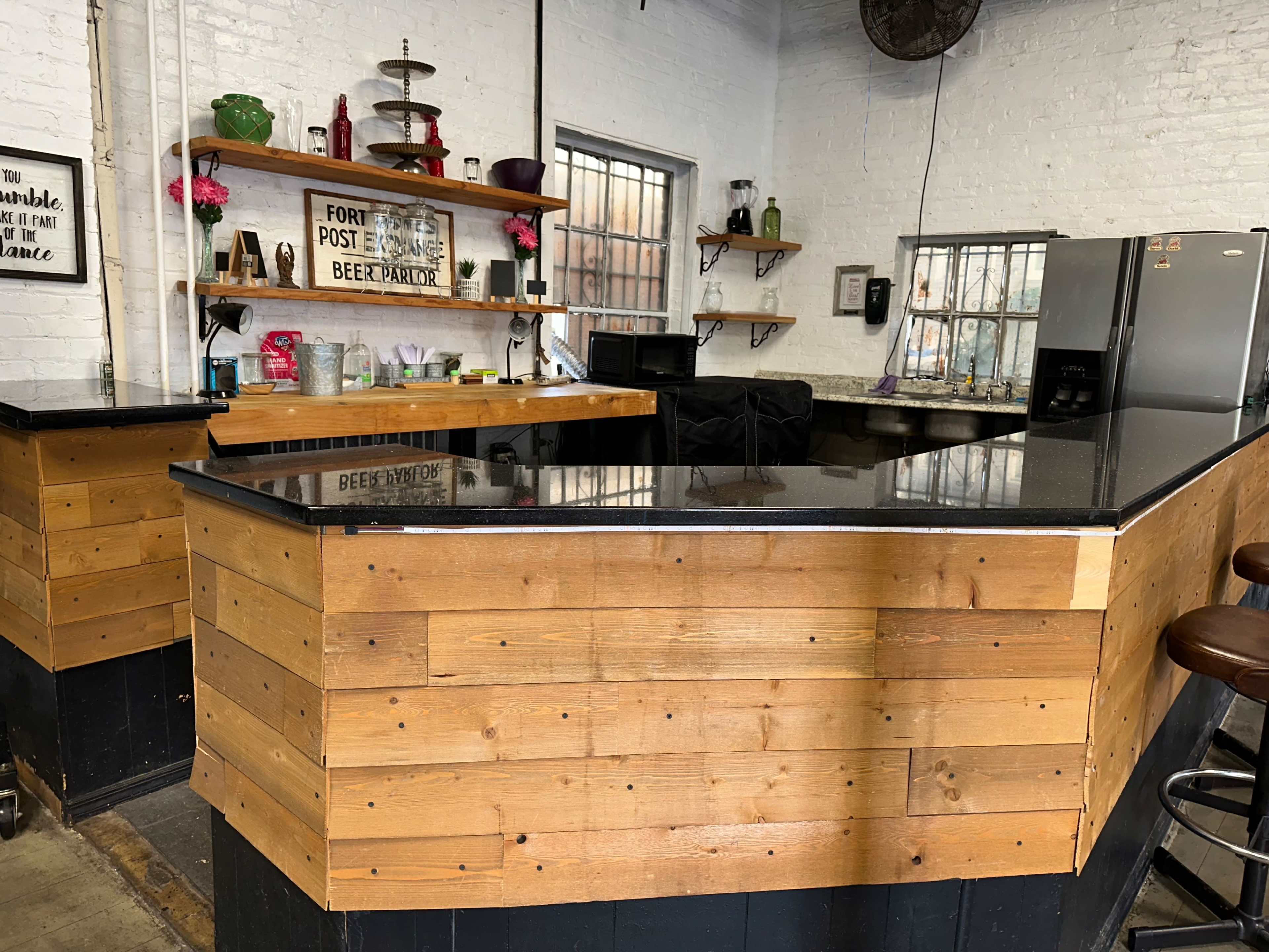 The image shows a wooden bar area with a black countertop, shelves displaying various items, and a refrigerator in a well-lit room with white brick walls.