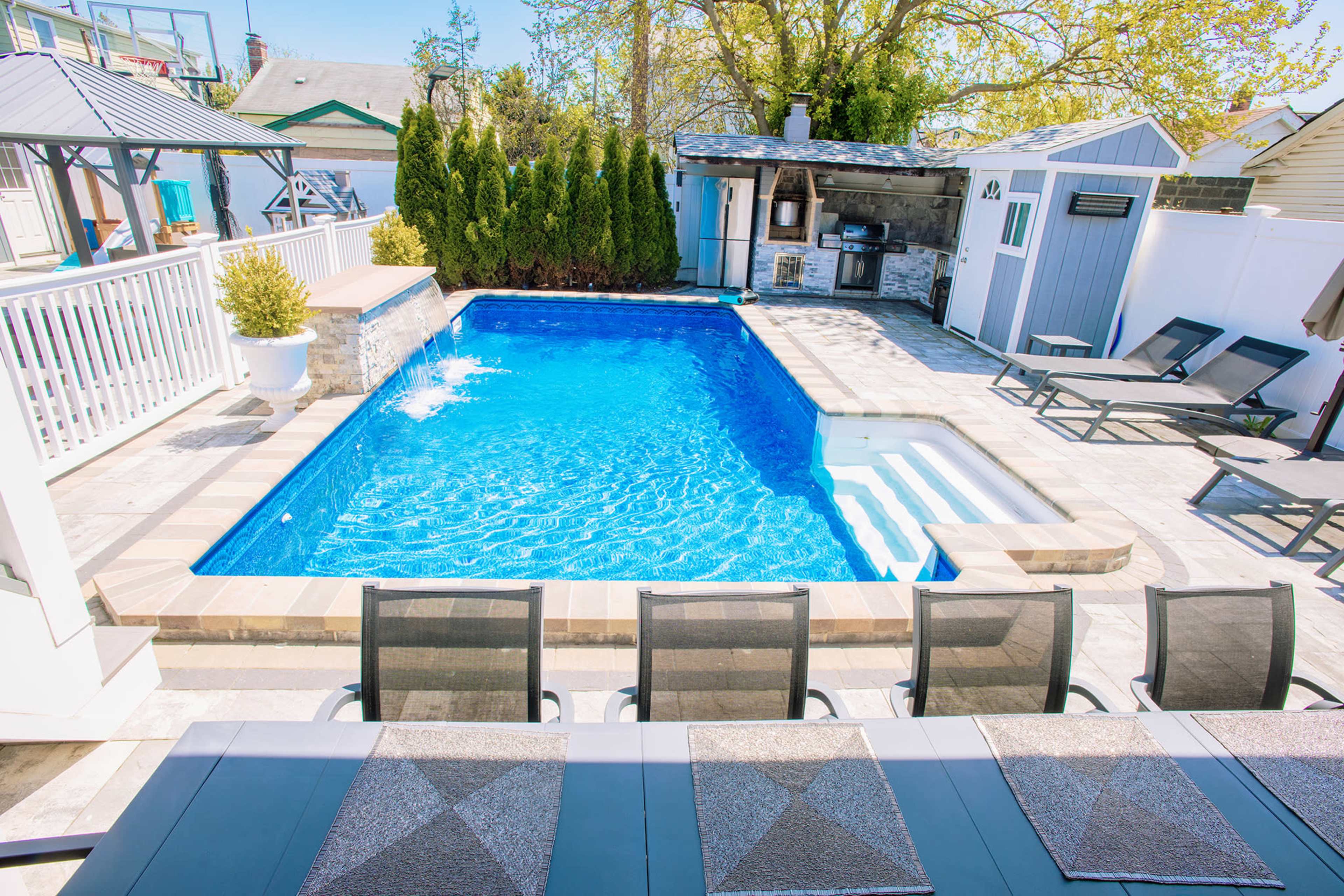 The image shows a backyard pool area featuring a blue swimming pool, a small hot tub, lounge chairs, and a barbecue grill setup with enclosed structures nearby.