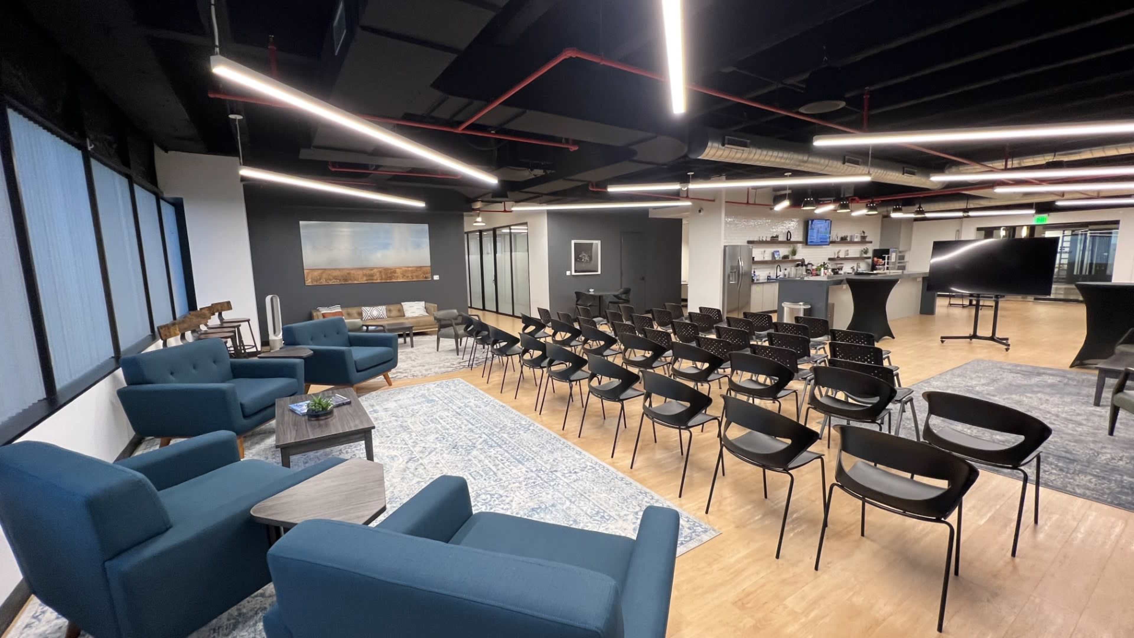 The image shows a modern office space with seating areas featuring blue chairs, a conference setup with black chairs facing a presentation area, and a kitchen area in the background.