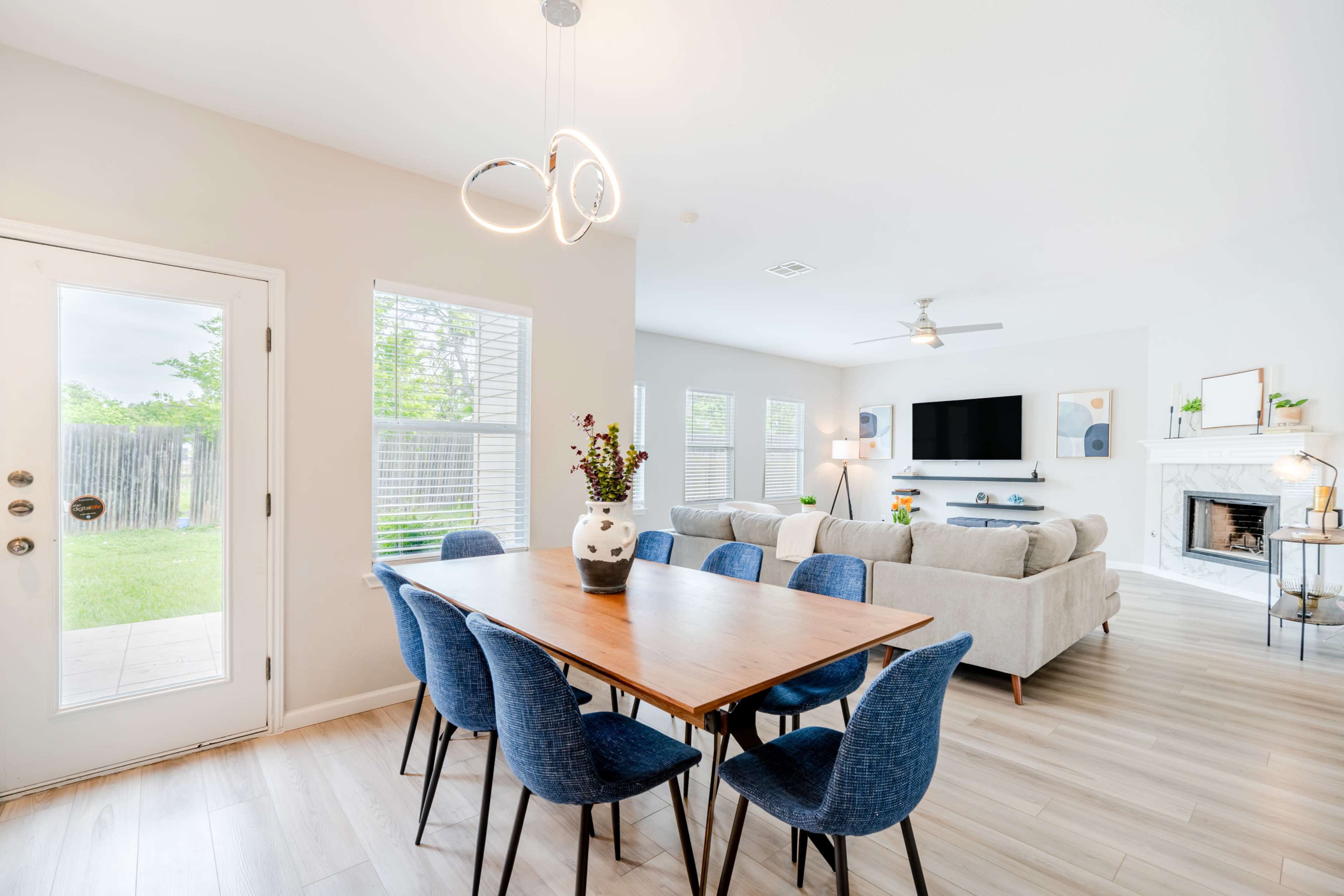 A dining area with a wooden table and blue chairs opens to a living space featuring a light-colored sofa and a wall-mounted television.