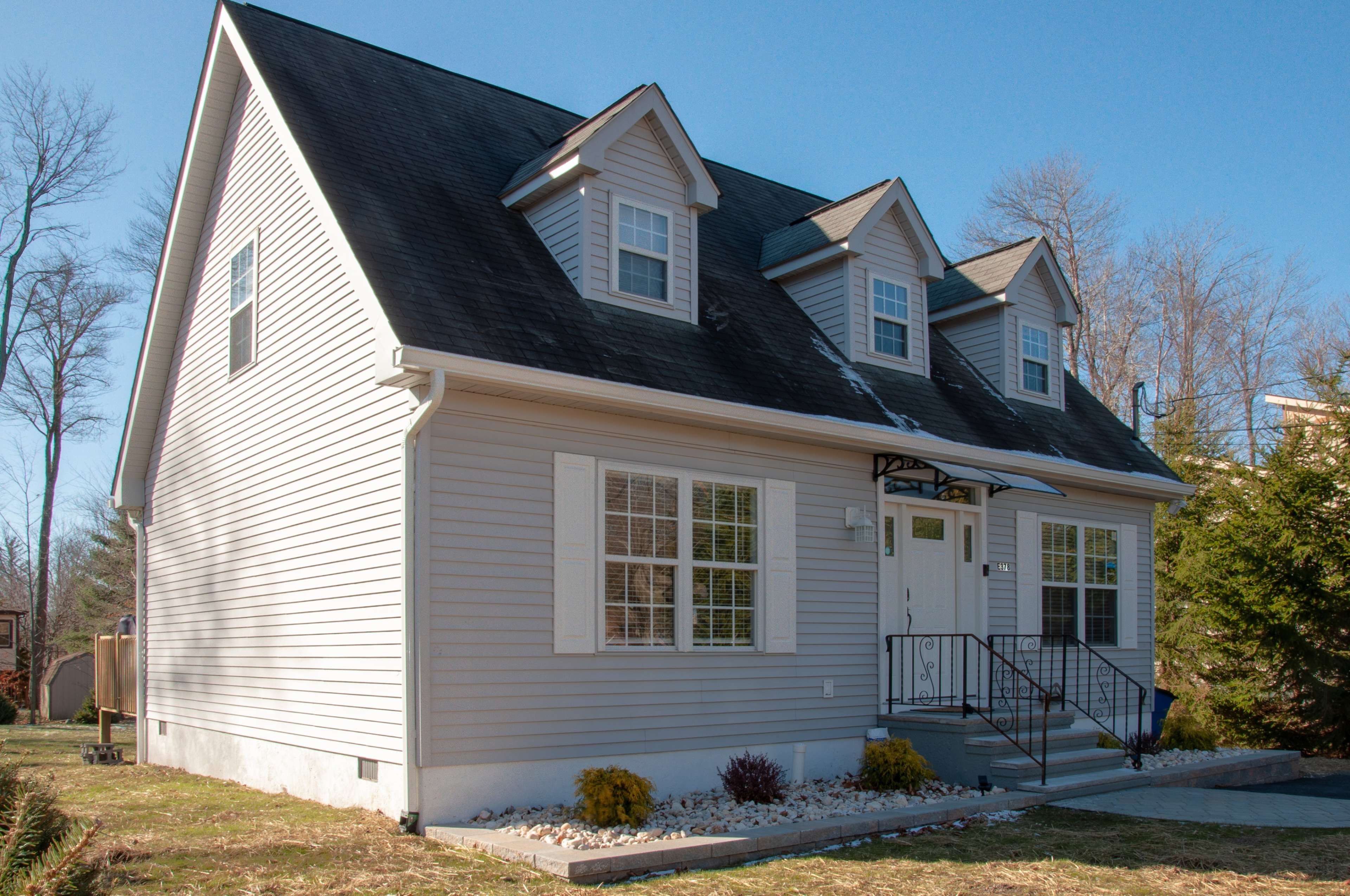 A light gray, two-story house features a sloped roof, dormer windows, and a neatly landscaped front yard.