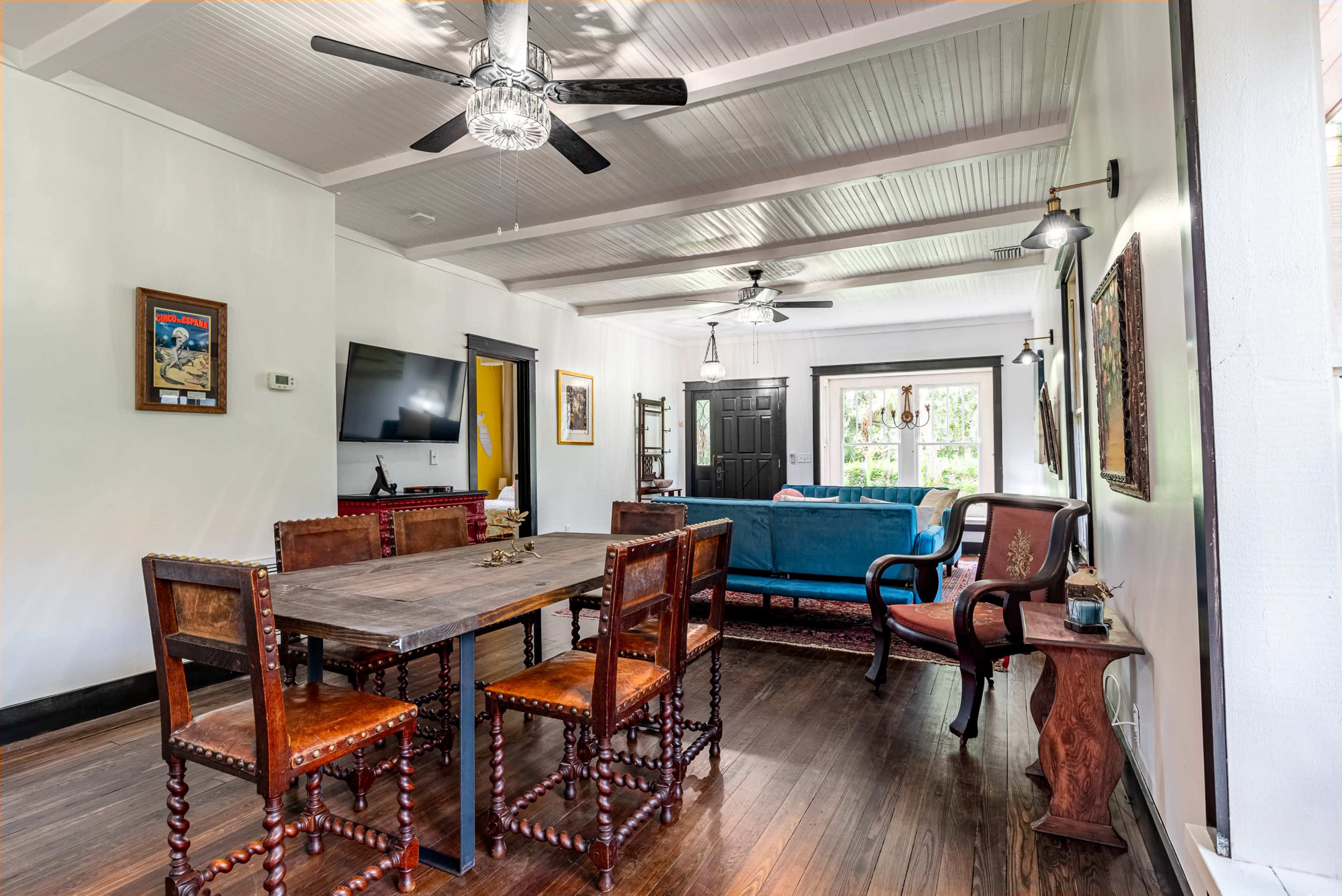 A living and dining area featuring a wooden dining table with chairs, a blue couch, and light fixtures, all under a ceiling with exposed beams.