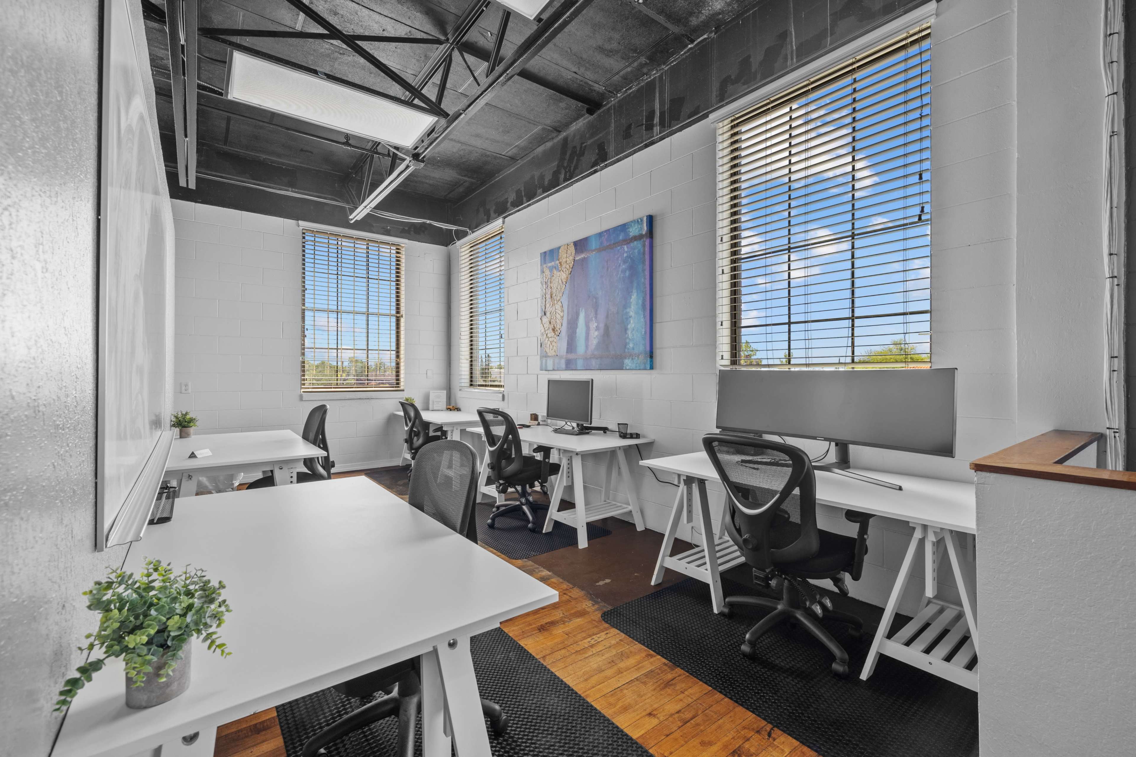 The image shows a modern office space with four workstations, large windows, and wooden flooring.
