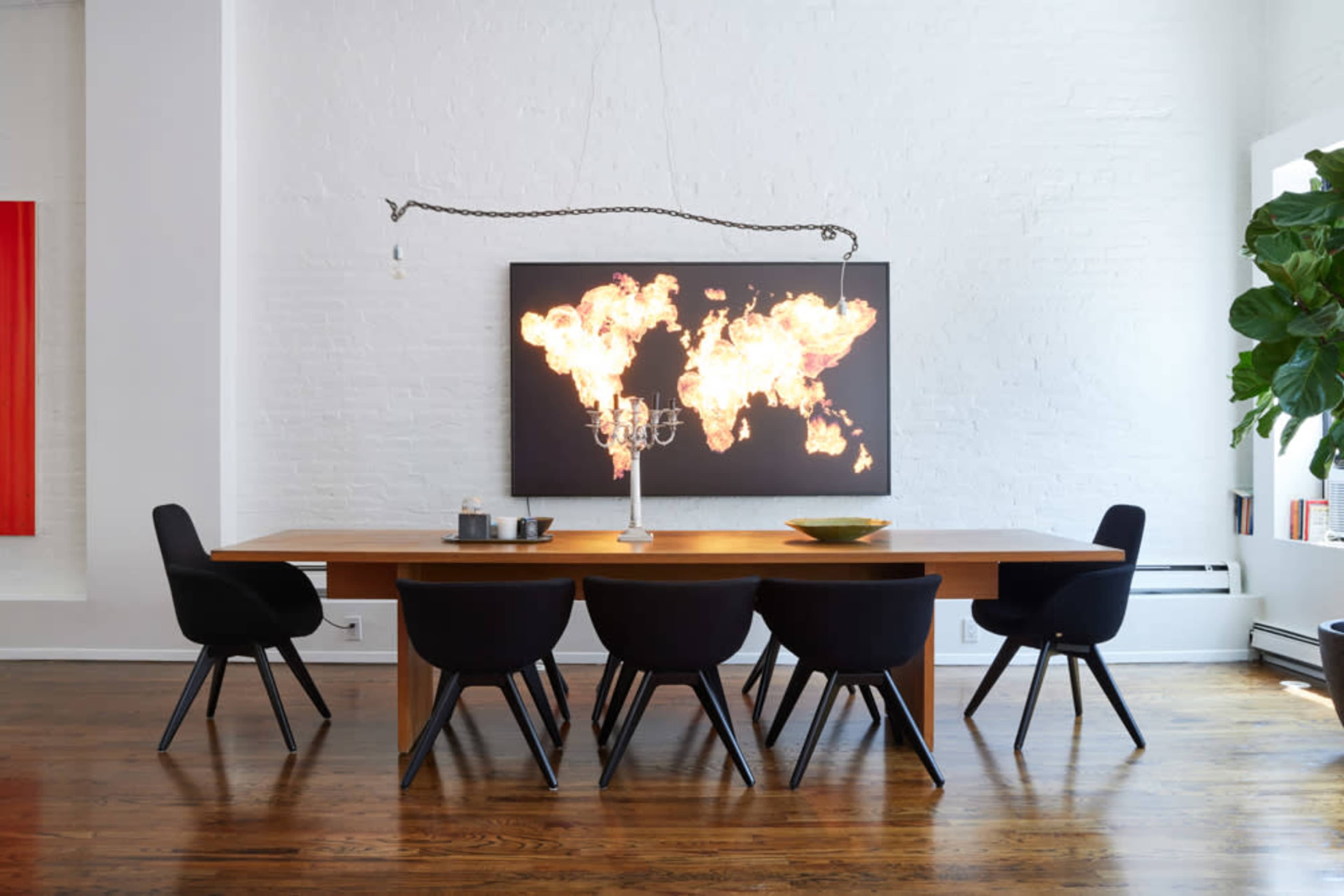 A long wooden dining table is set with a centerpiece and candles, surrounded by six black chairs, beneath a large artwork depicting a flaming world map.