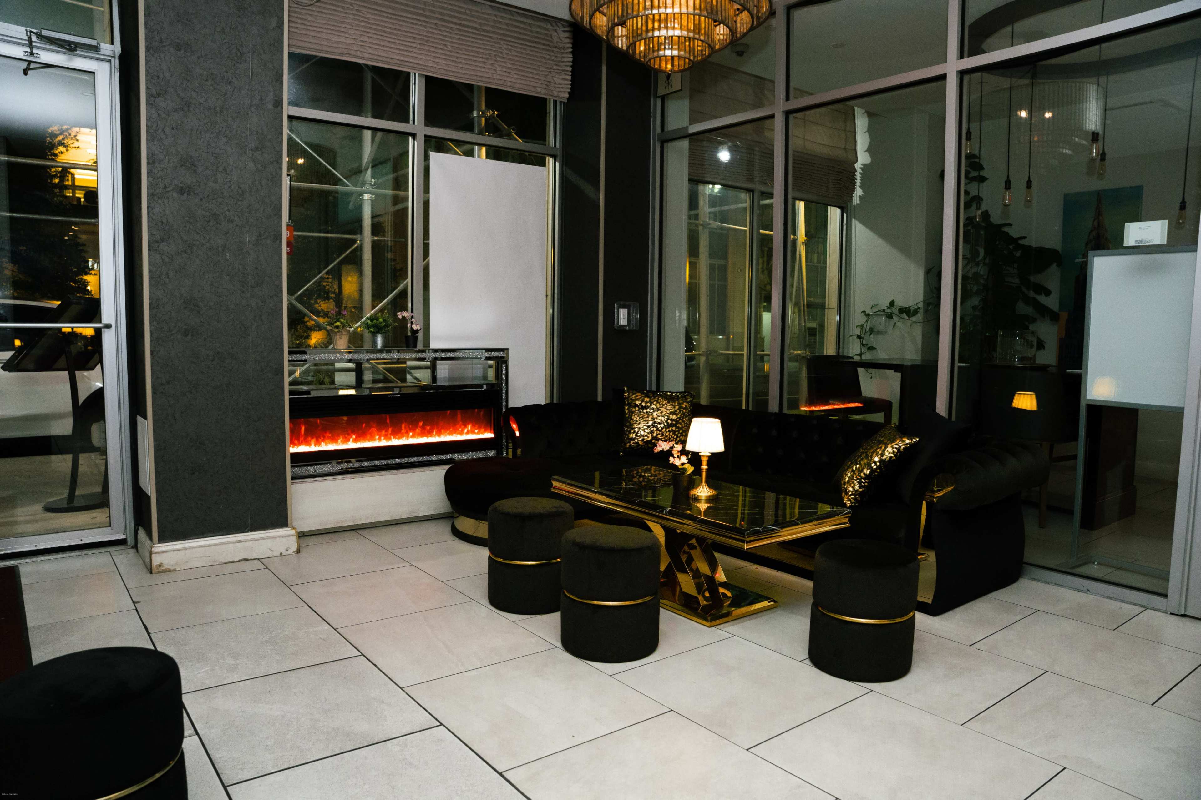 The image shows a modern lounge area with a black sofa, golden accents, a glass coffee table, and an electric fireplace.
