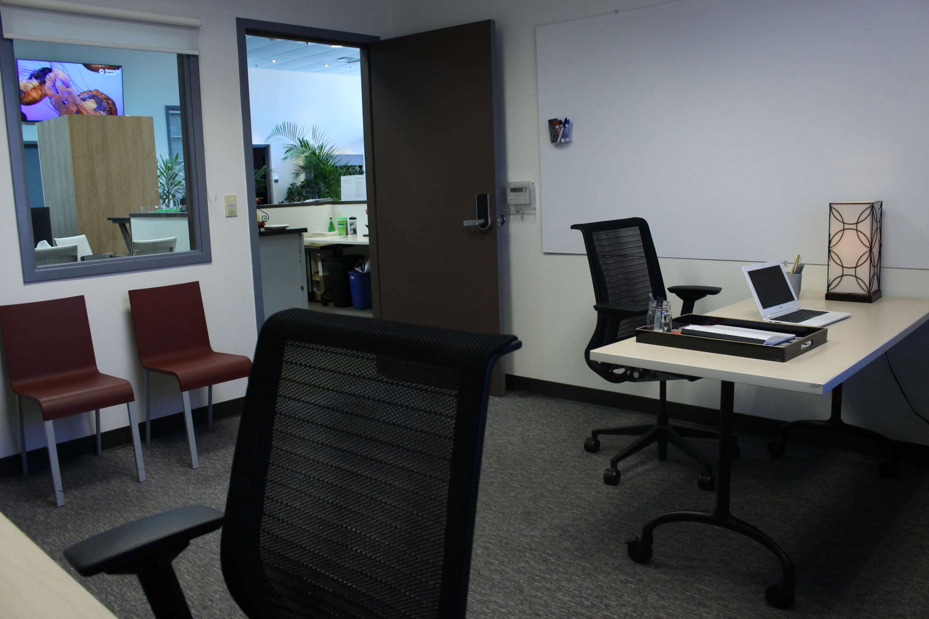 A modern office space with a desk, laptop, and two red chairs positioned against a wall, while a large whiteboard is visible nearby.