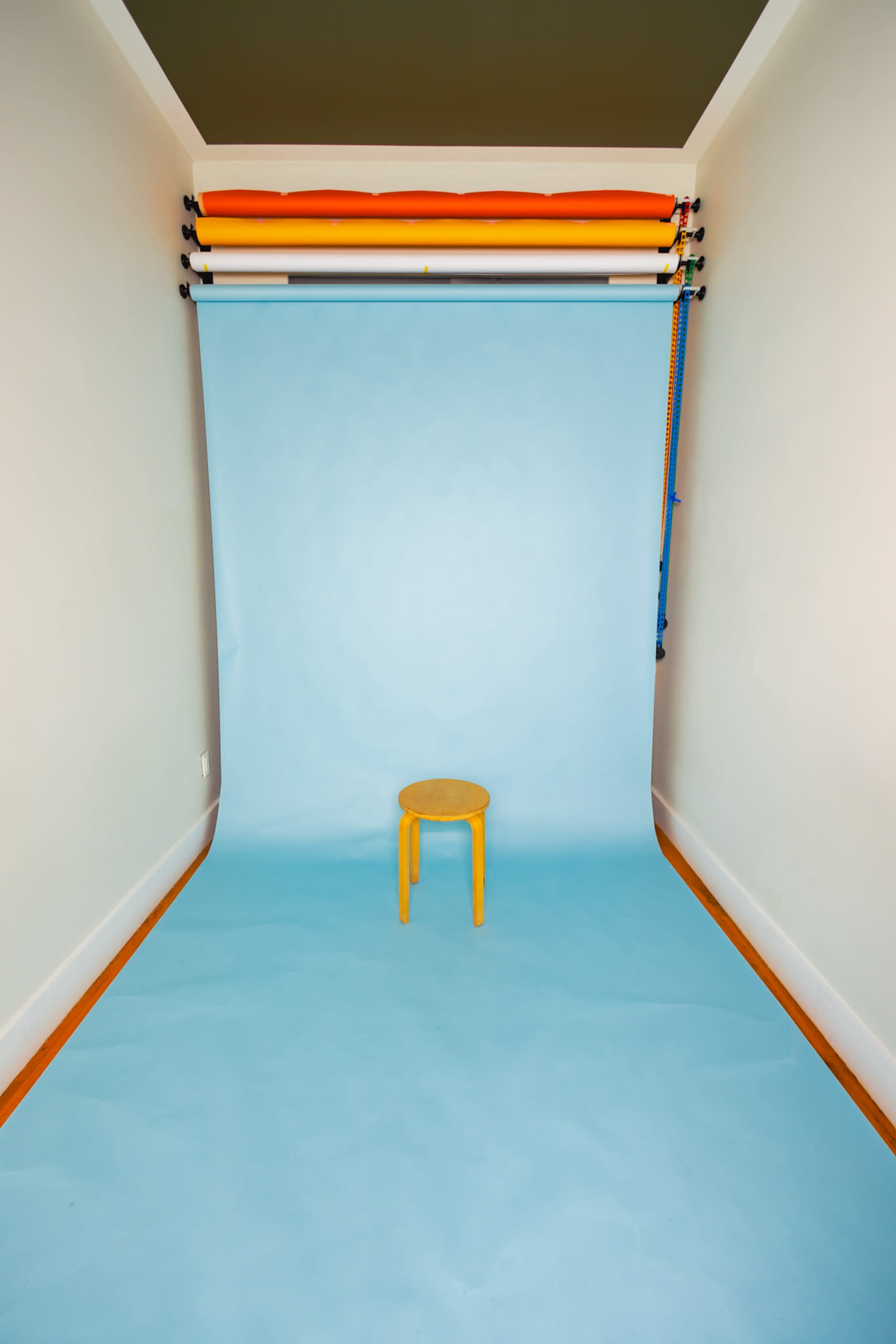 A simple studio space features a wooden stool positioned on light blue flooring, with a backdrop of blue paper and colored support poles above.