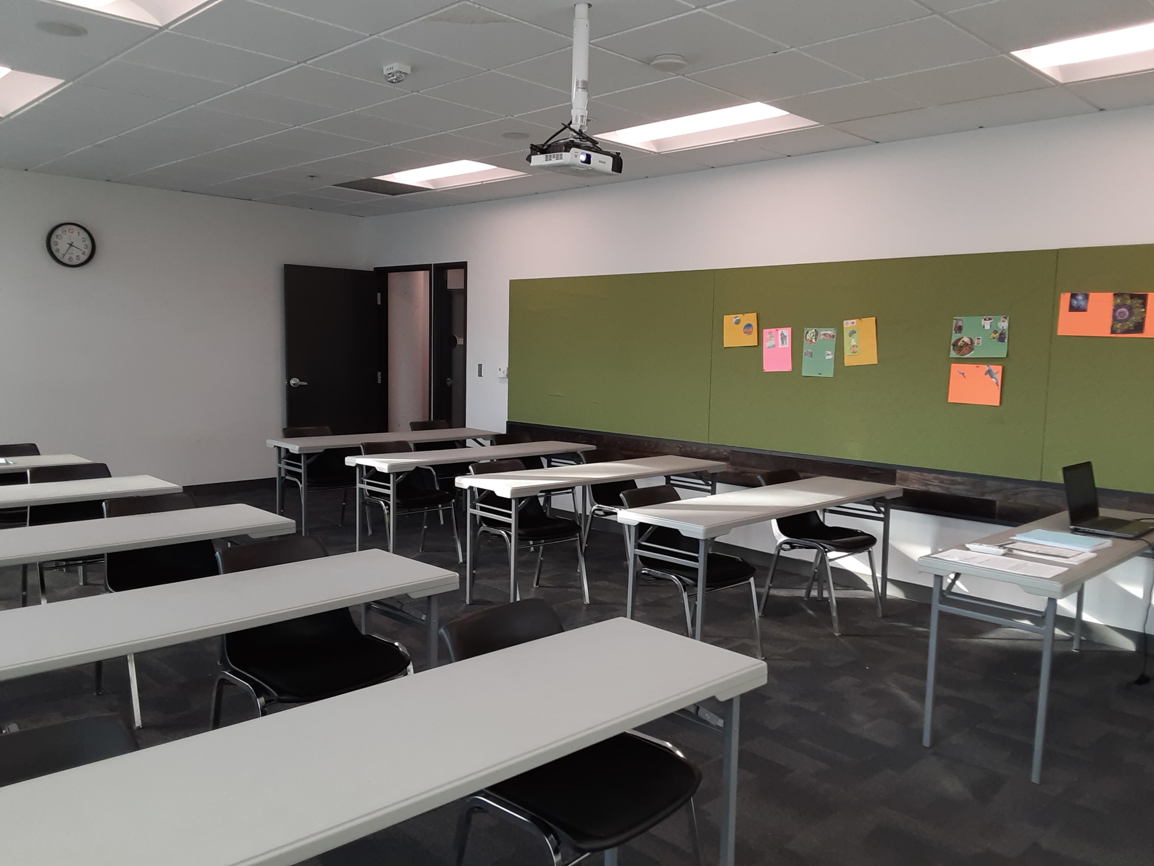 A classroom is shown with multiple rows of white tables and black chairs, a clock on the wall, and a green bulletin board displaying colorful posters.