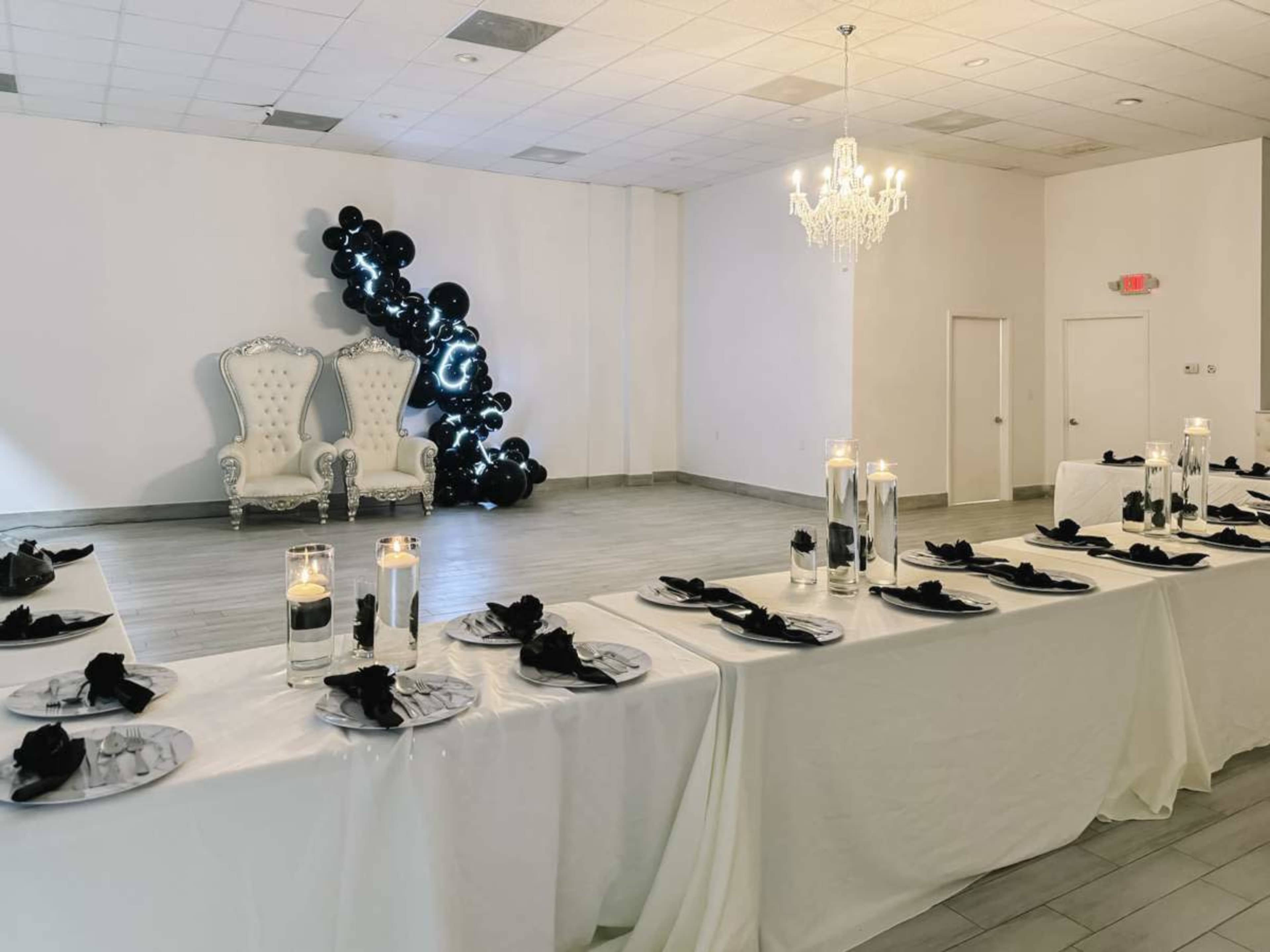 The image shows a beautifully arranged banquet hall with a long table set for a formal event, featuring white tablecloths, black napkins, and a decorative backdrop of balloons, alongside an ornate white throne chair and a chandelier.