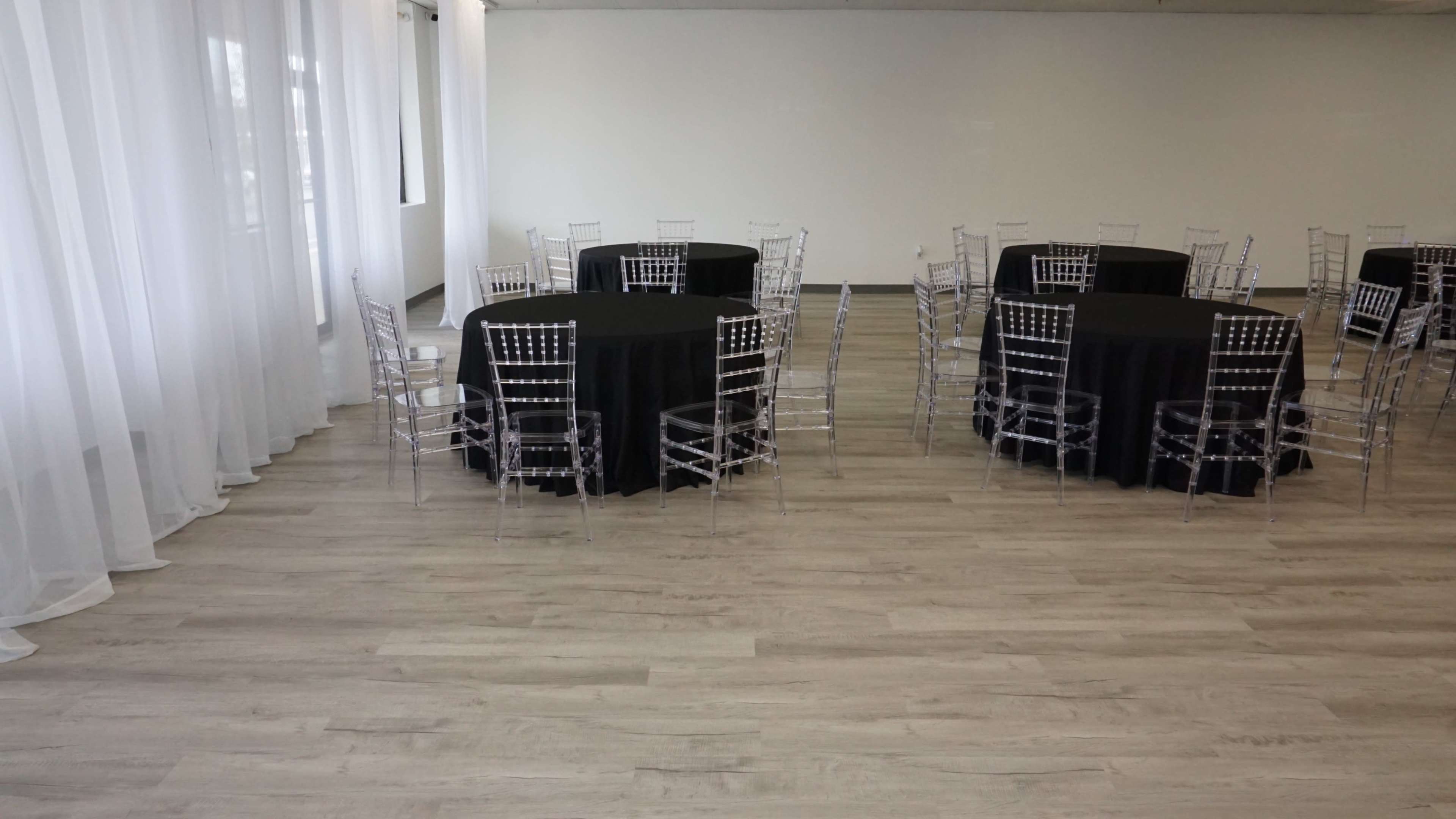 The image shows a spacious room with multiple round tables covered with black tablecloths, surrounded by clear plastic chairs.