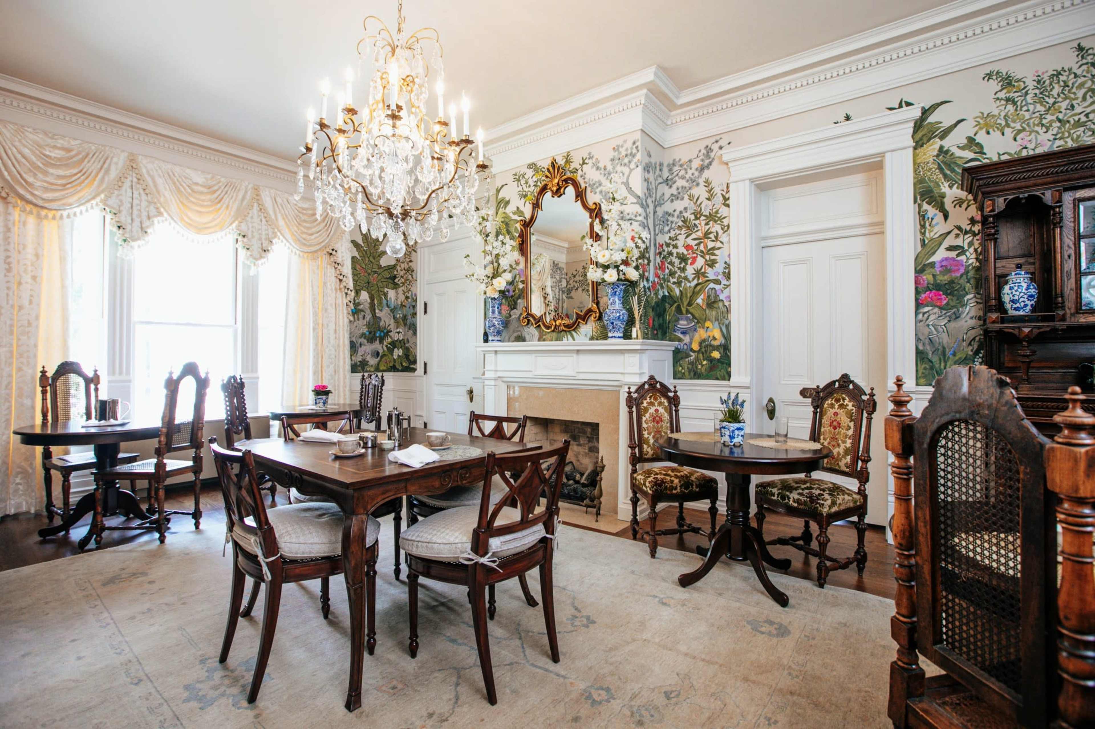 The image shows an elegantly decorated dining room with floral wallpaper, a chandelier, and wooden furniture arranged around a dining table.