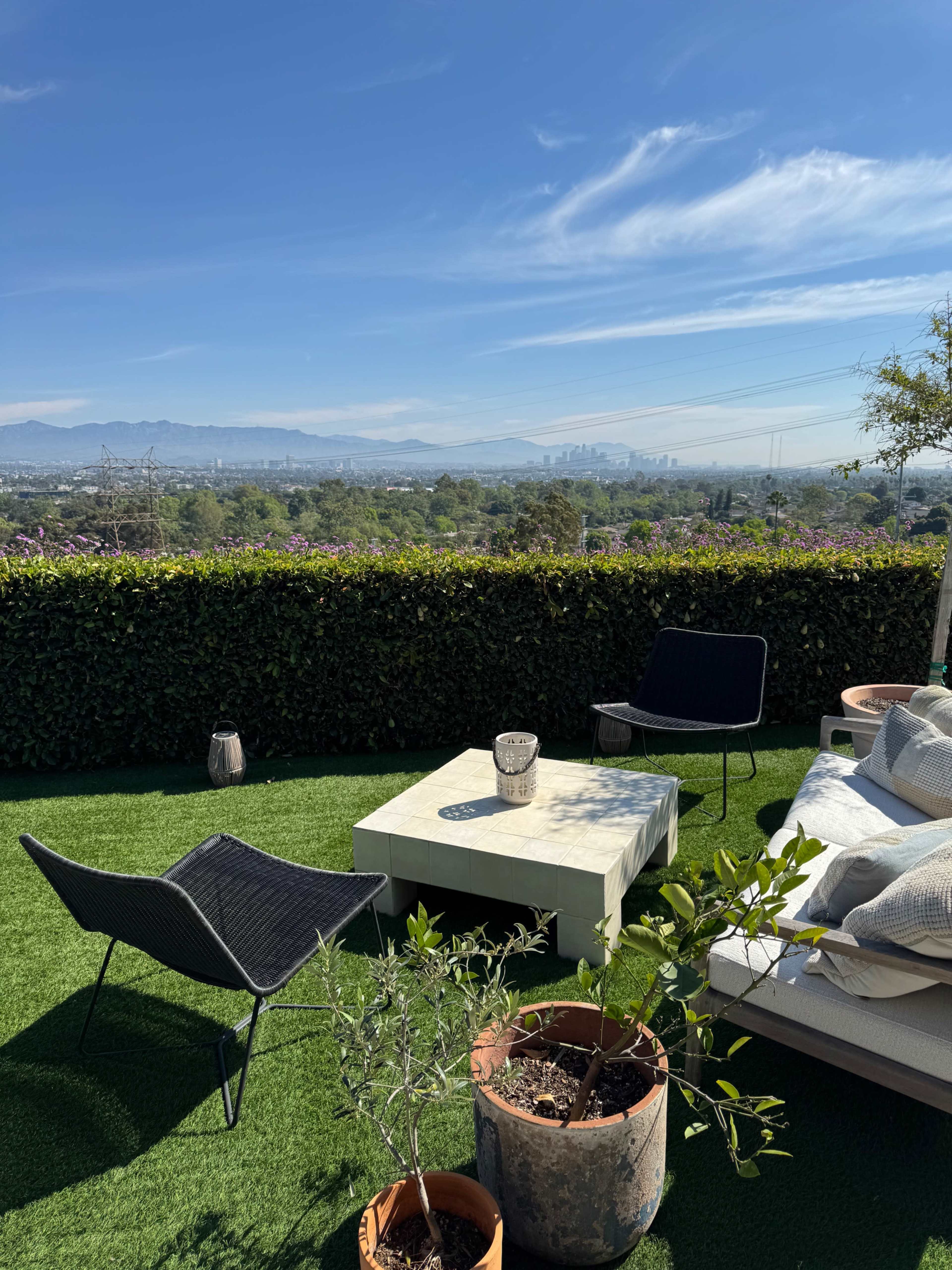 The image shows a landscaped terrace with a white coffee table, two black chairs, potted plants, and a view of a city skyline and mountains under a clear blue sky.