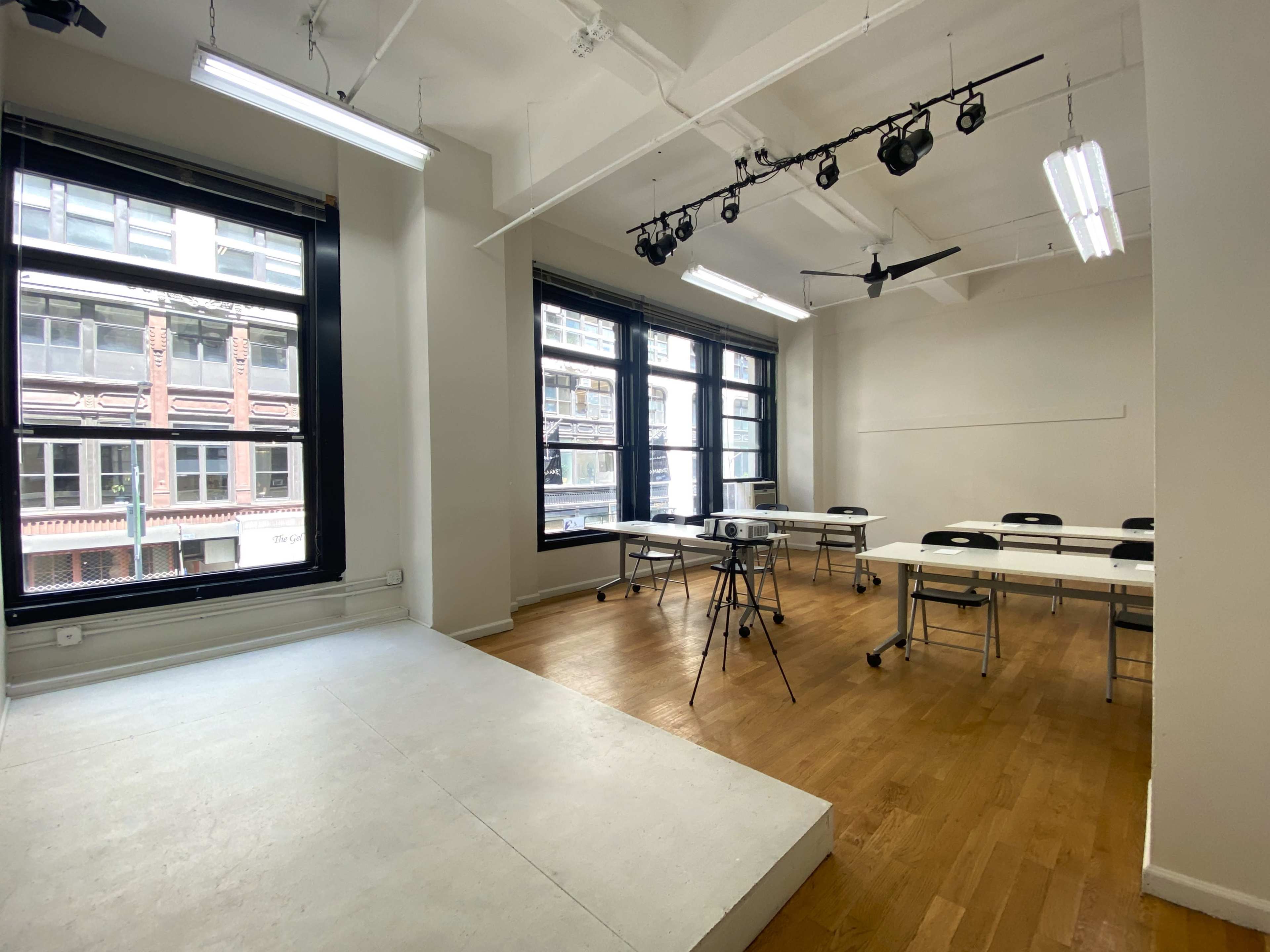 A bright, empty classroom features a projector setup, several tables with chairs, and large windows overlooking a city street.