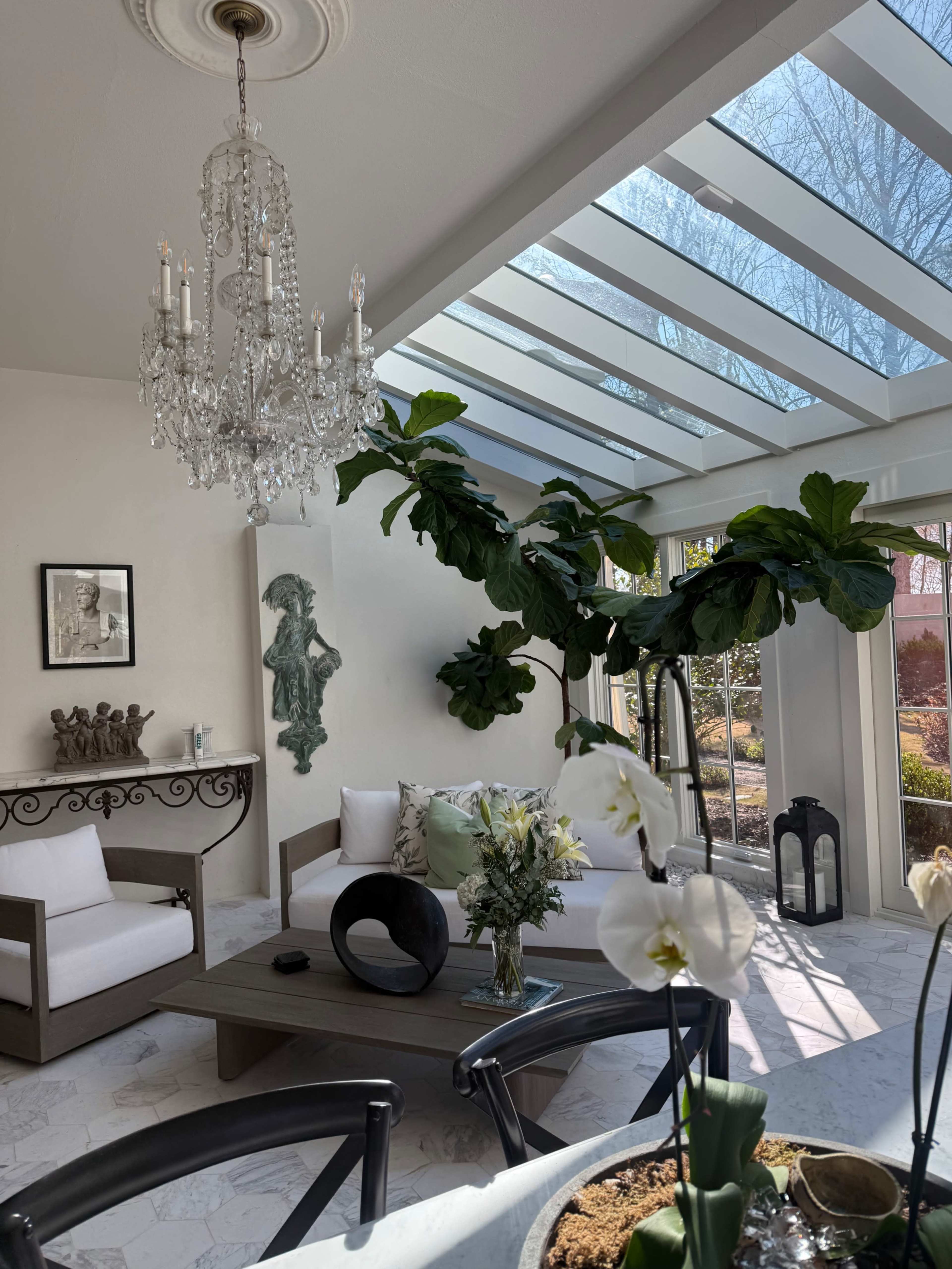 A bright living room features a modern chandelier, large potted plants, and a sunlit area with a view of the outdoors.