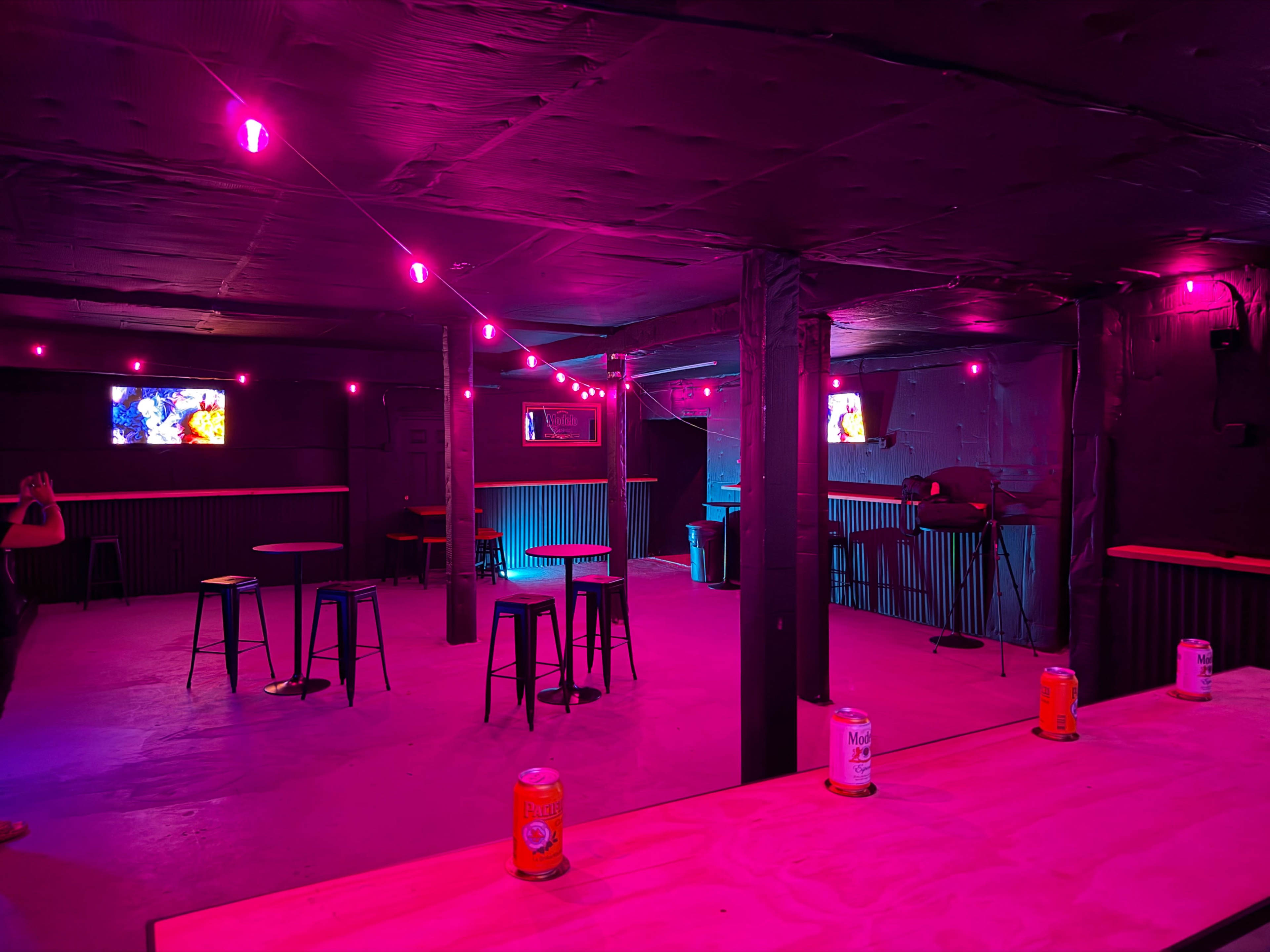 A dimly-lit bar space features pink lighting, empty stools, and a few cans on a table.