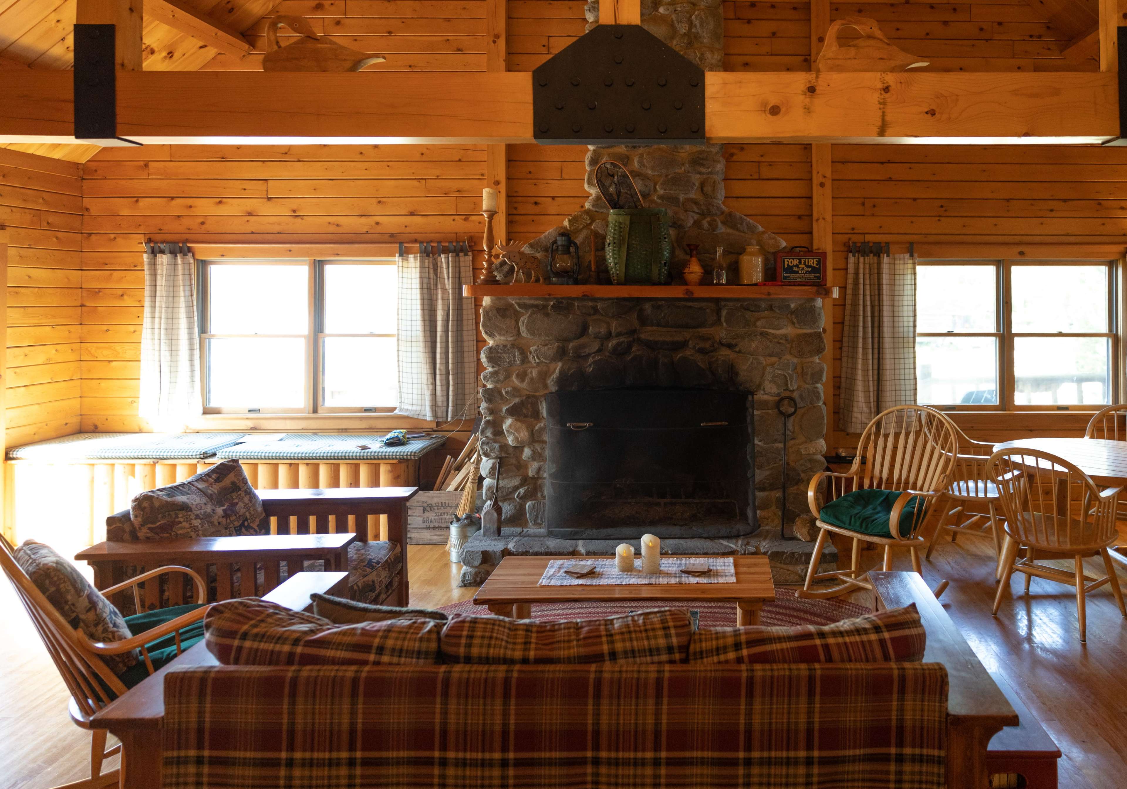 The interior of a wooden cabin features a stone fireplace, a plaid sofa, and a dining area with wooden chairs.