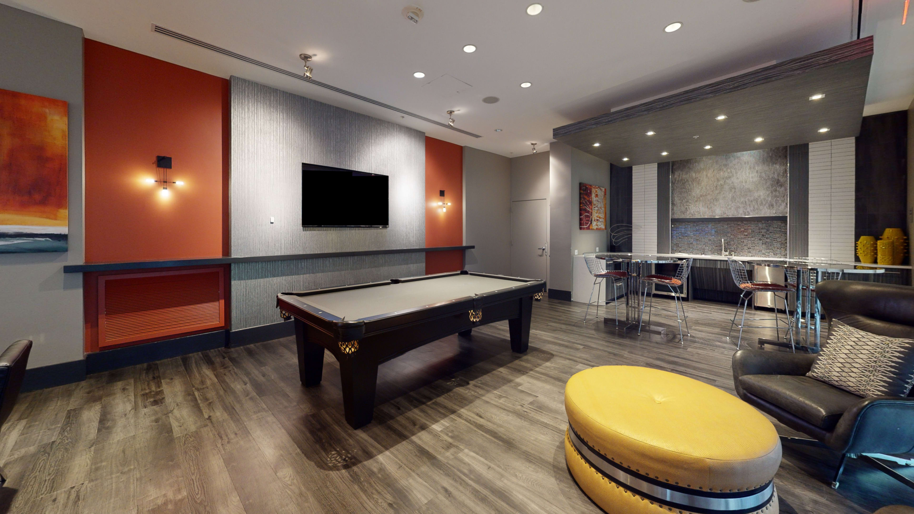A modern lounge area featuring a billiards table, a television mounted on the wall, and a bar with a high countertop and seating.