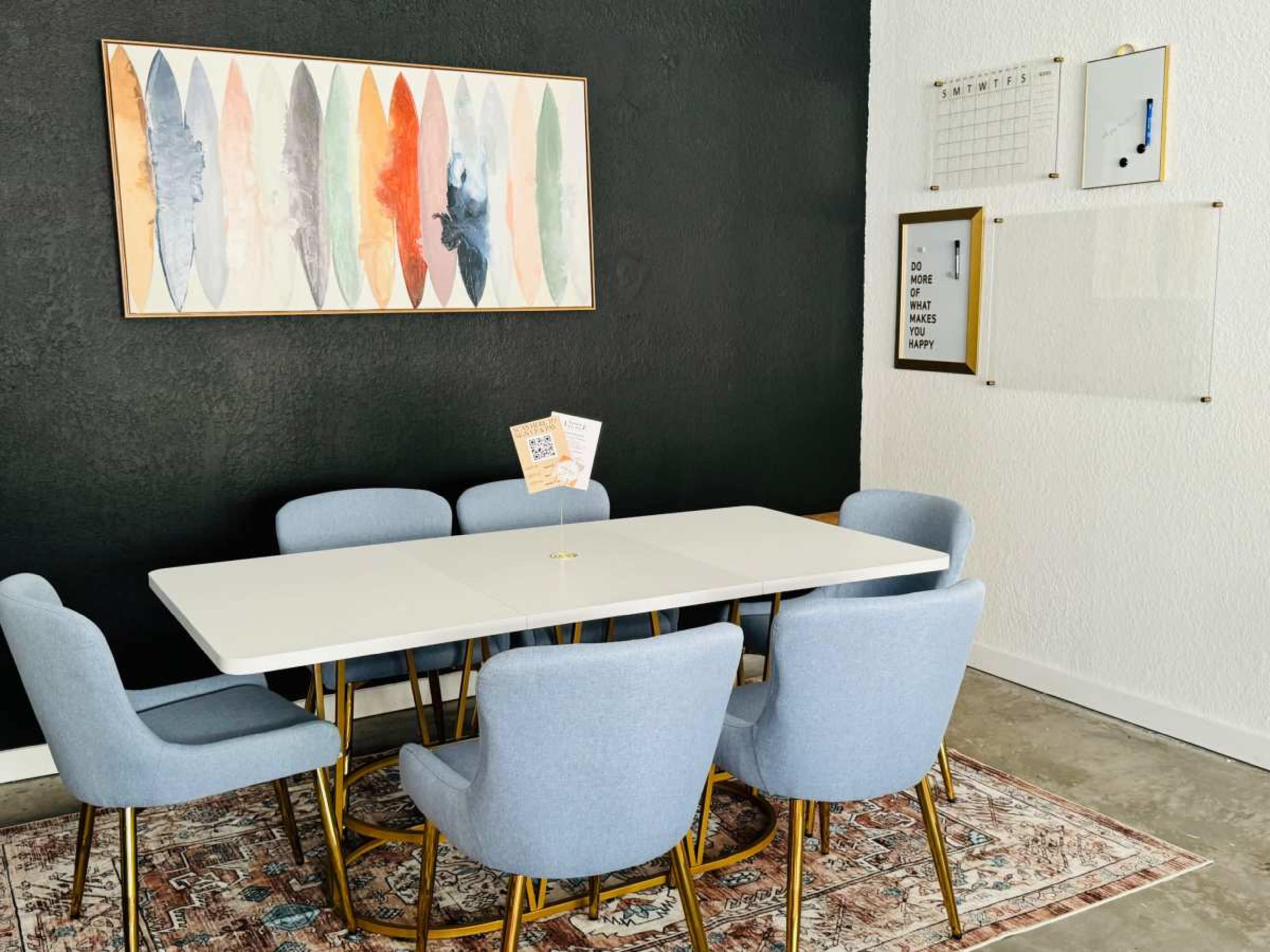 A modern dining area features a white table surrounded by six light blue chairs on a patterned rug, with artwork and a calendar on the wall.