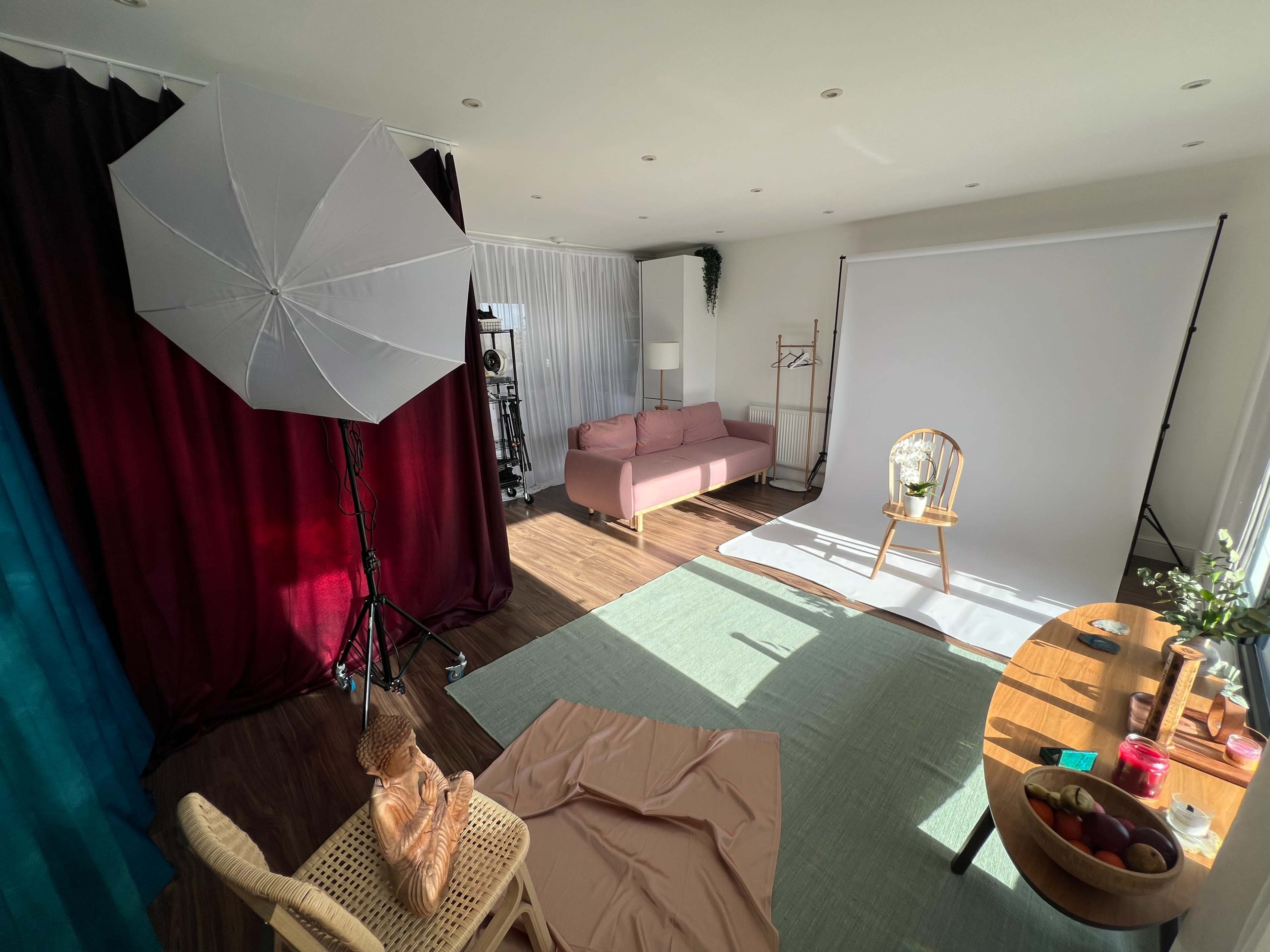 The image shows a well-lit photography studio featuring a backdrop, a chair with a plant, a sofa, and some props arranged around a table.