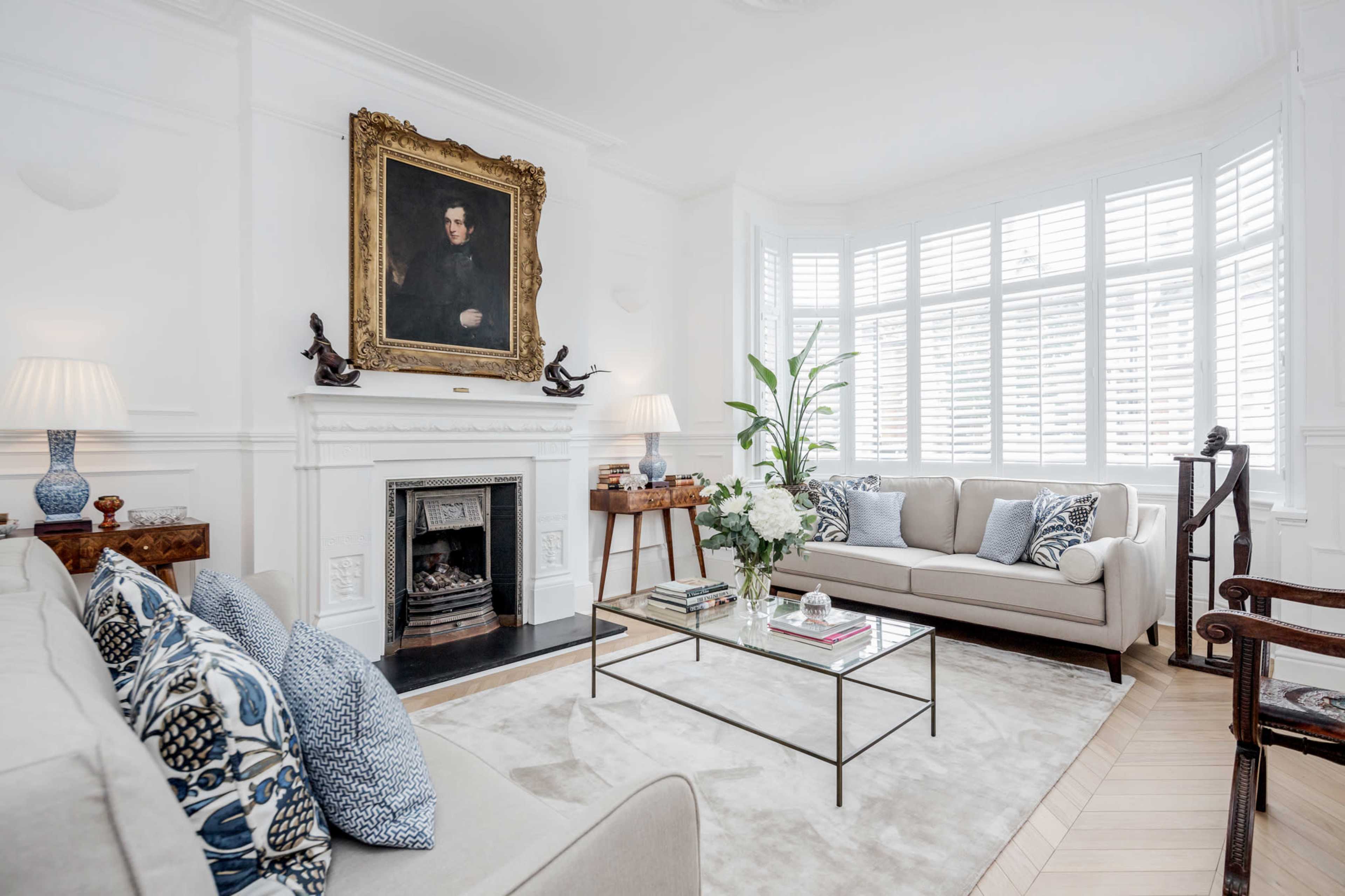A bright living room features a fireplace, a large portrait above the mantel, a plush sofa with decorative pillows, and a glass coffee table surrounded by elegant decor.