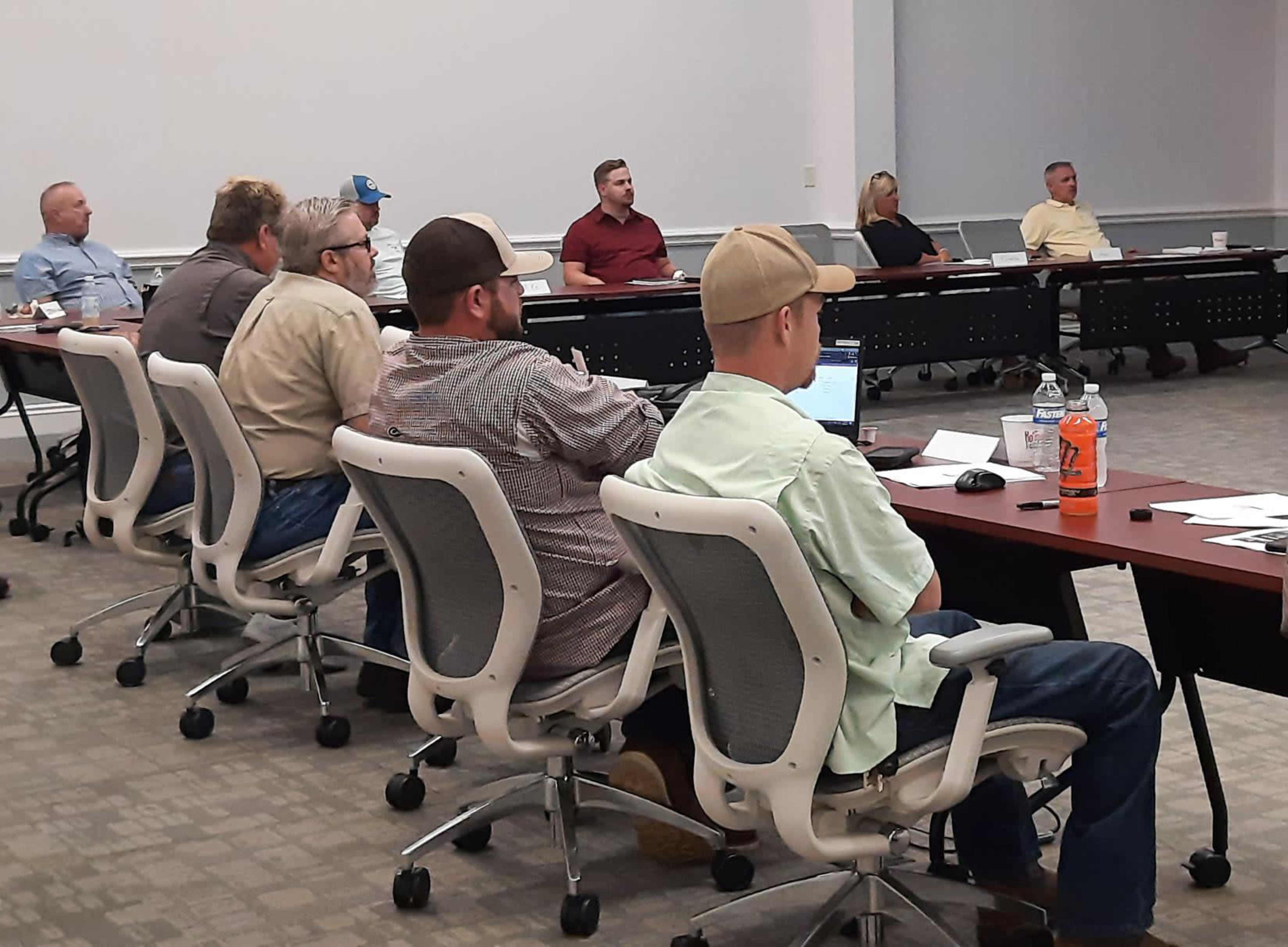 A group of people is seated in a conference room, attentively facing a presentation at the front.