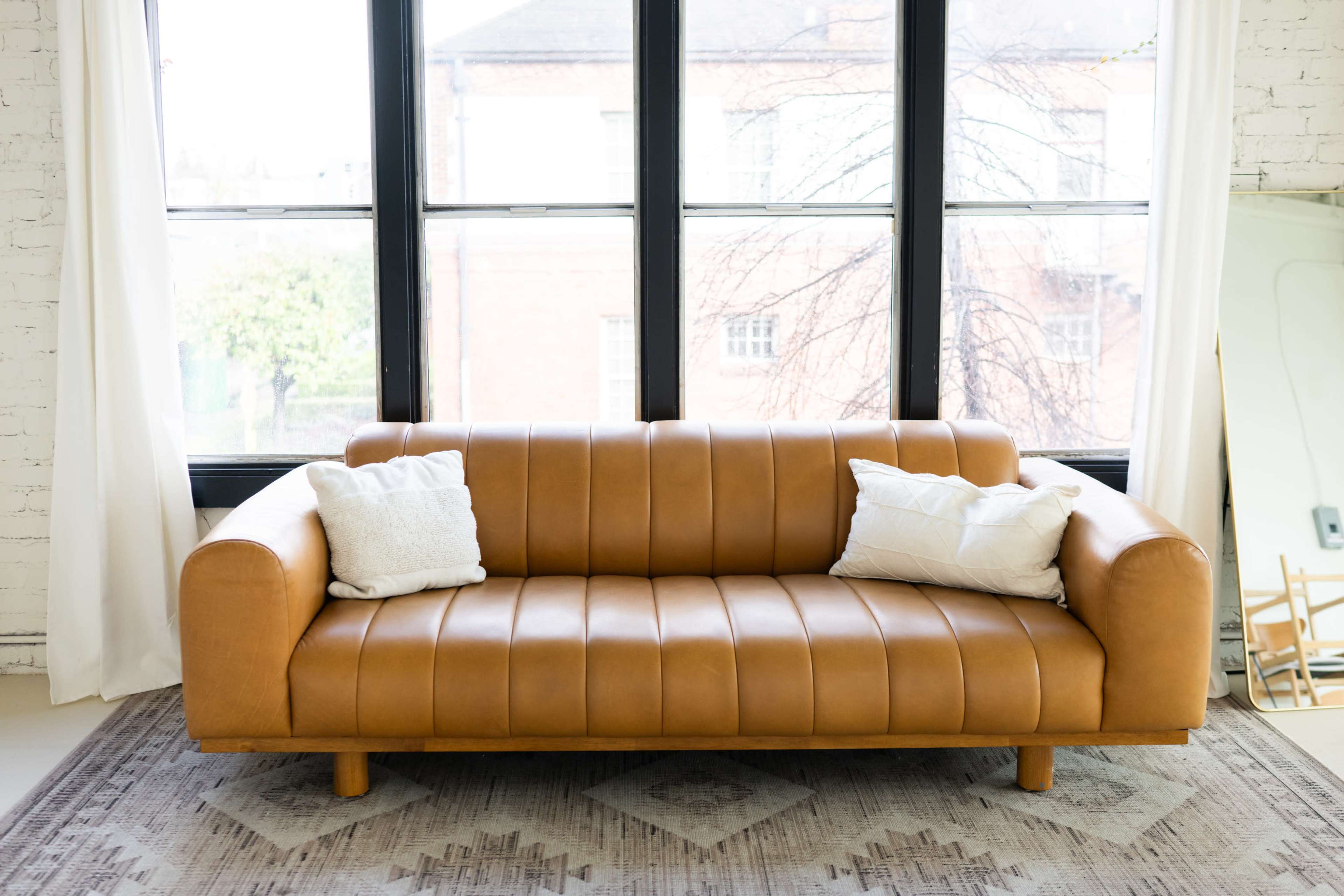 A brown leather sofa with vertical stitching and two white throw pillows is positioned in front of large windows.