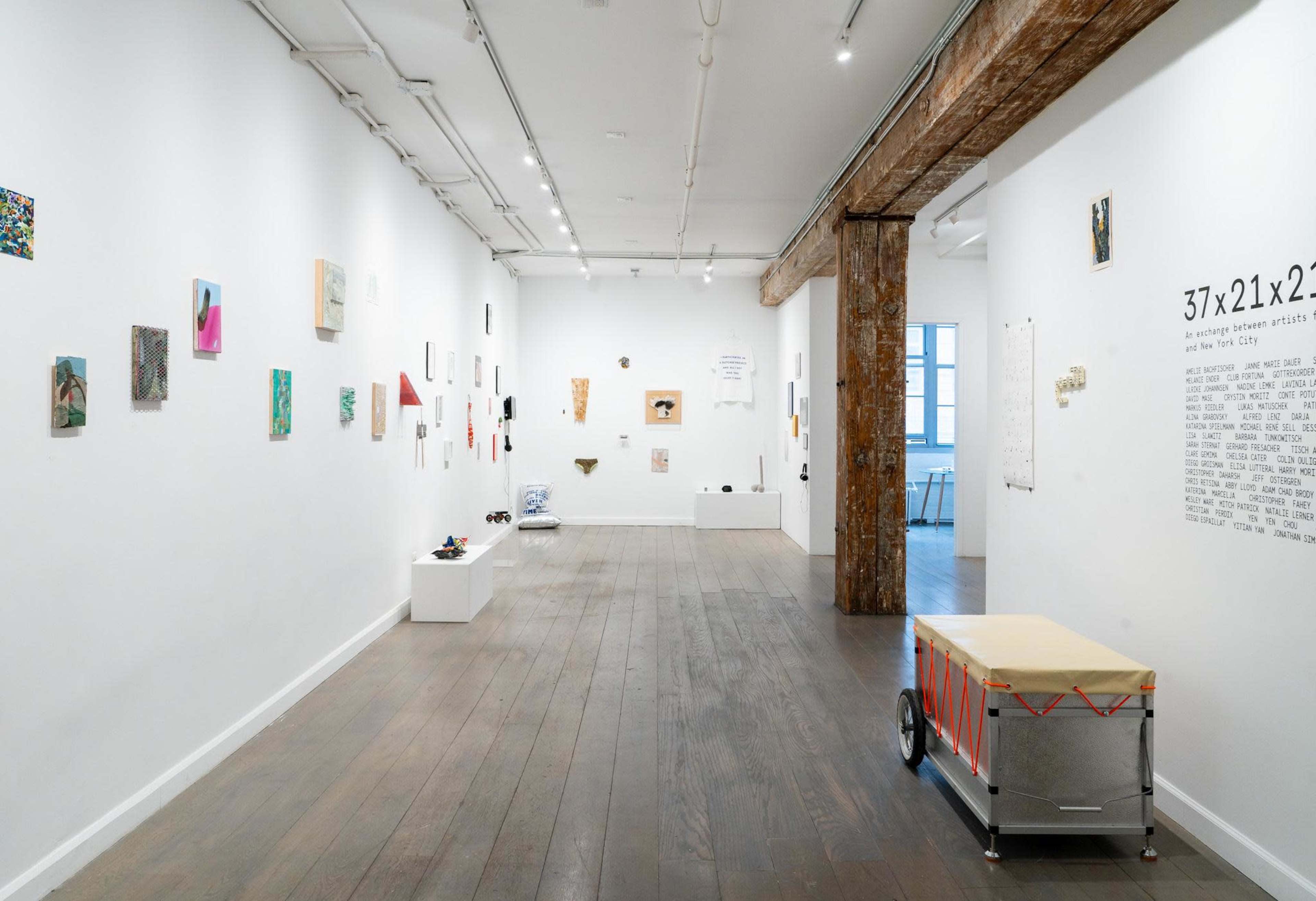 The image shows a contemporary art gallery featuring white walls adorned with various artworks and a wooden support beam on the right.