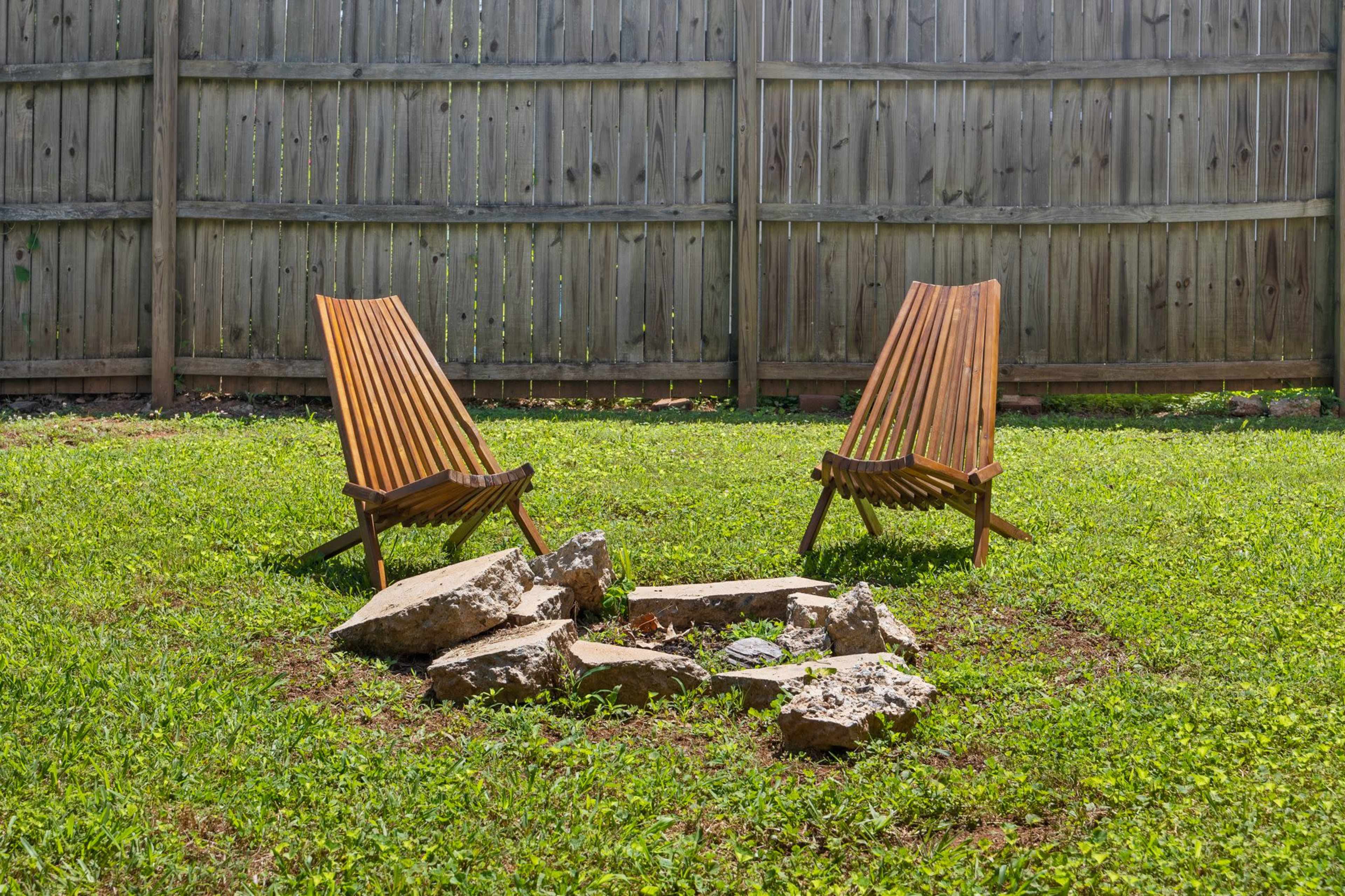 Two wooden lounge chairs are positioned on a grassy lawn facing a small fire pit made of stones, in a backyard enclosed by a wooden fence.