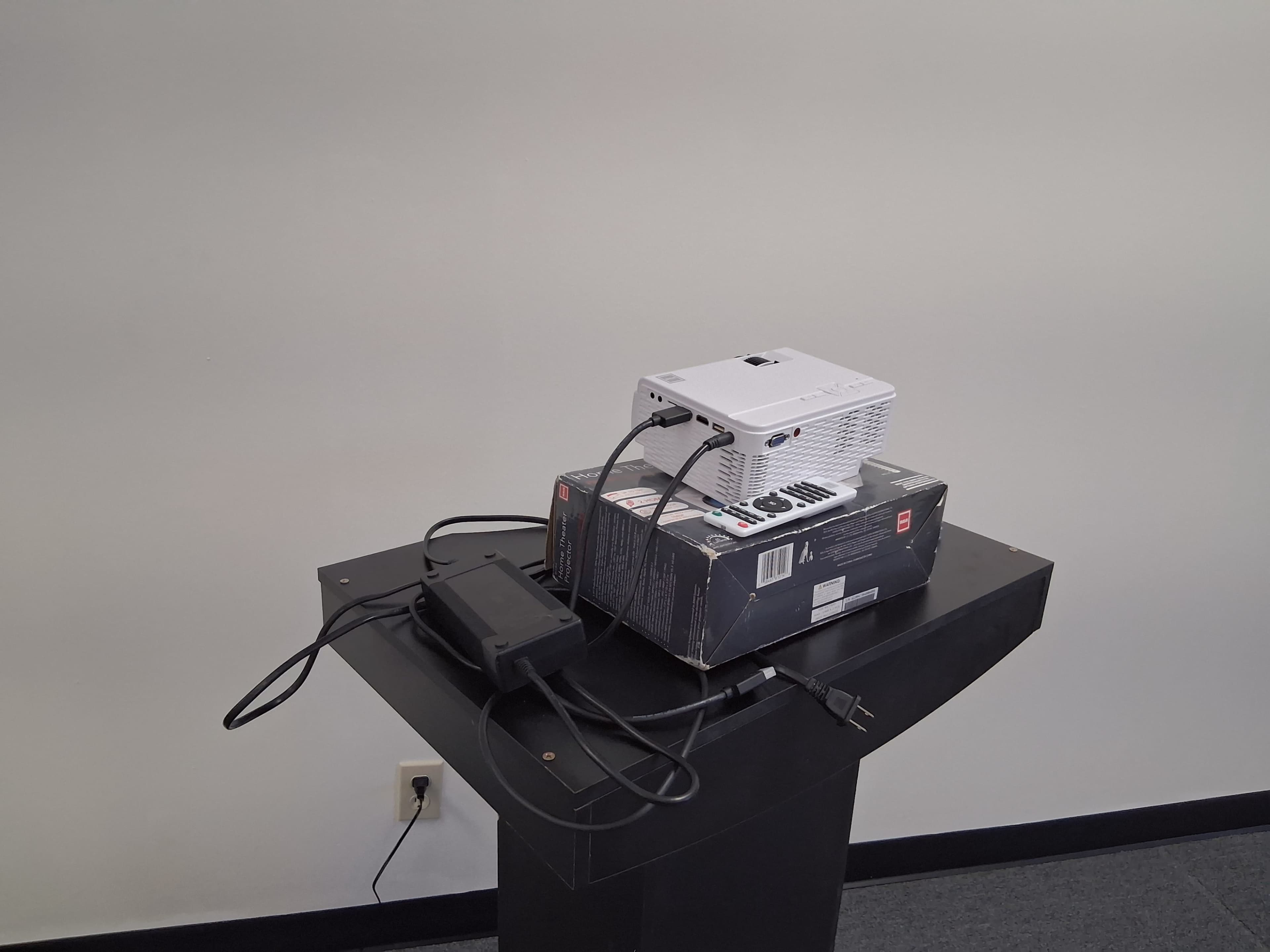 A small projector is placed on top of a box, with various cables connected to it, sitting on a black podium against a light-colored wall.