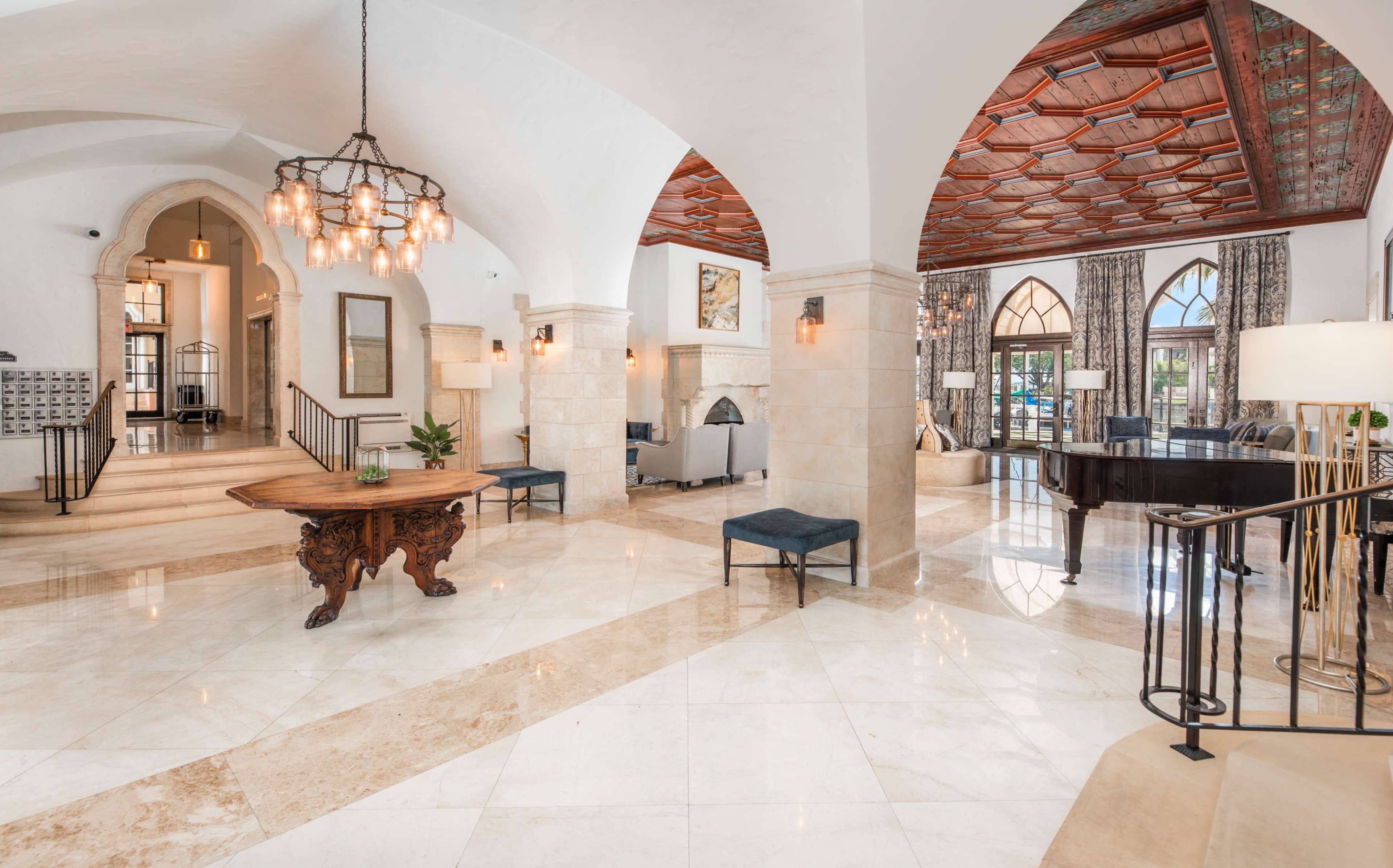 A spacious, elegantly designed lobby featuring high ceilings, marble flooring, and decorative arches, with a piano and seating areas arranged around a central table.
